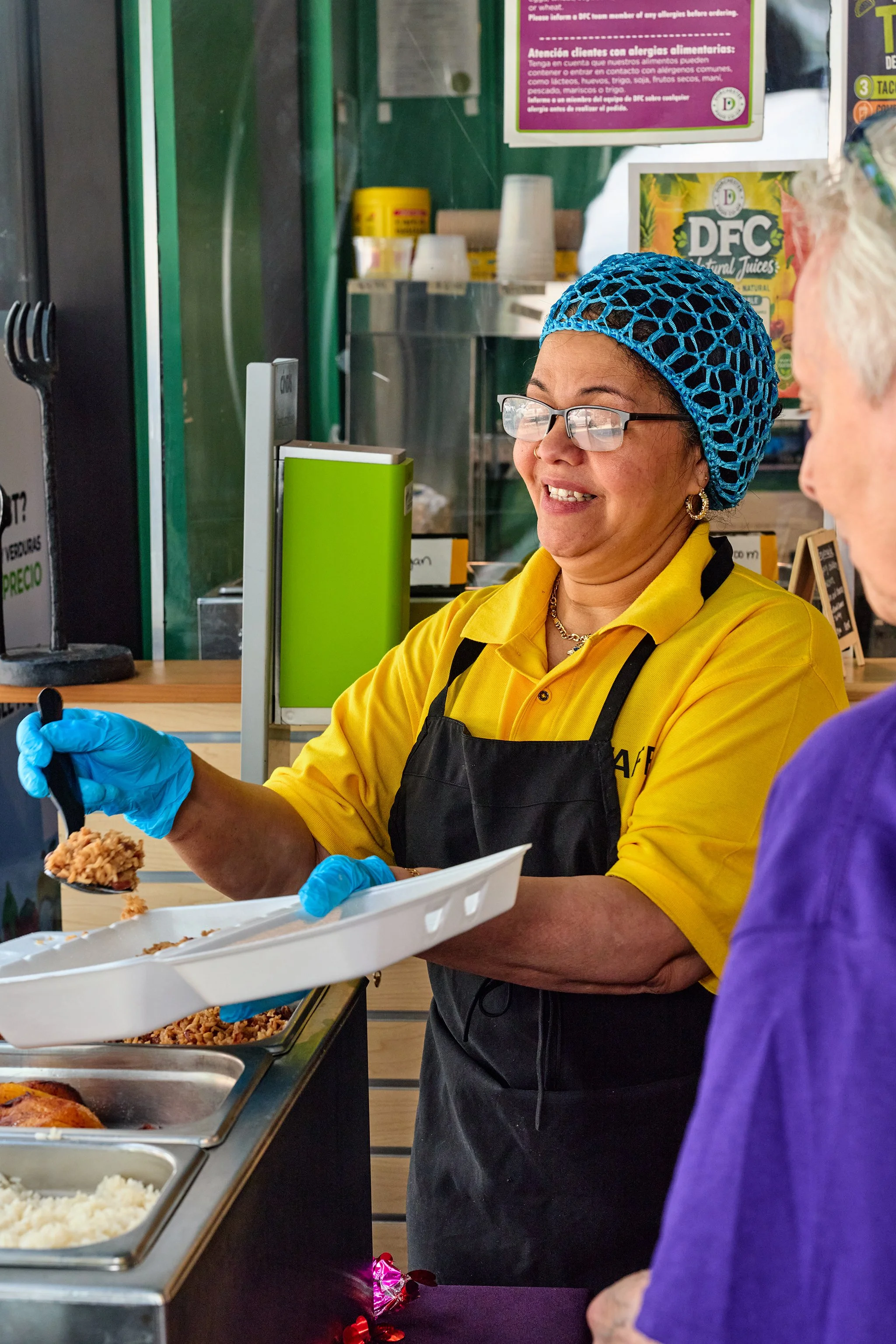 A woman wearing a yellow uniform, glasses, and a blue netted hair cover is serving food from a cafeteria-style serving counter. She is using tongs to scoop food onto a white tray, with various food trays visible in front of her. There are signs and posters in the background and another person in a purple shirt is partially visible to the right.