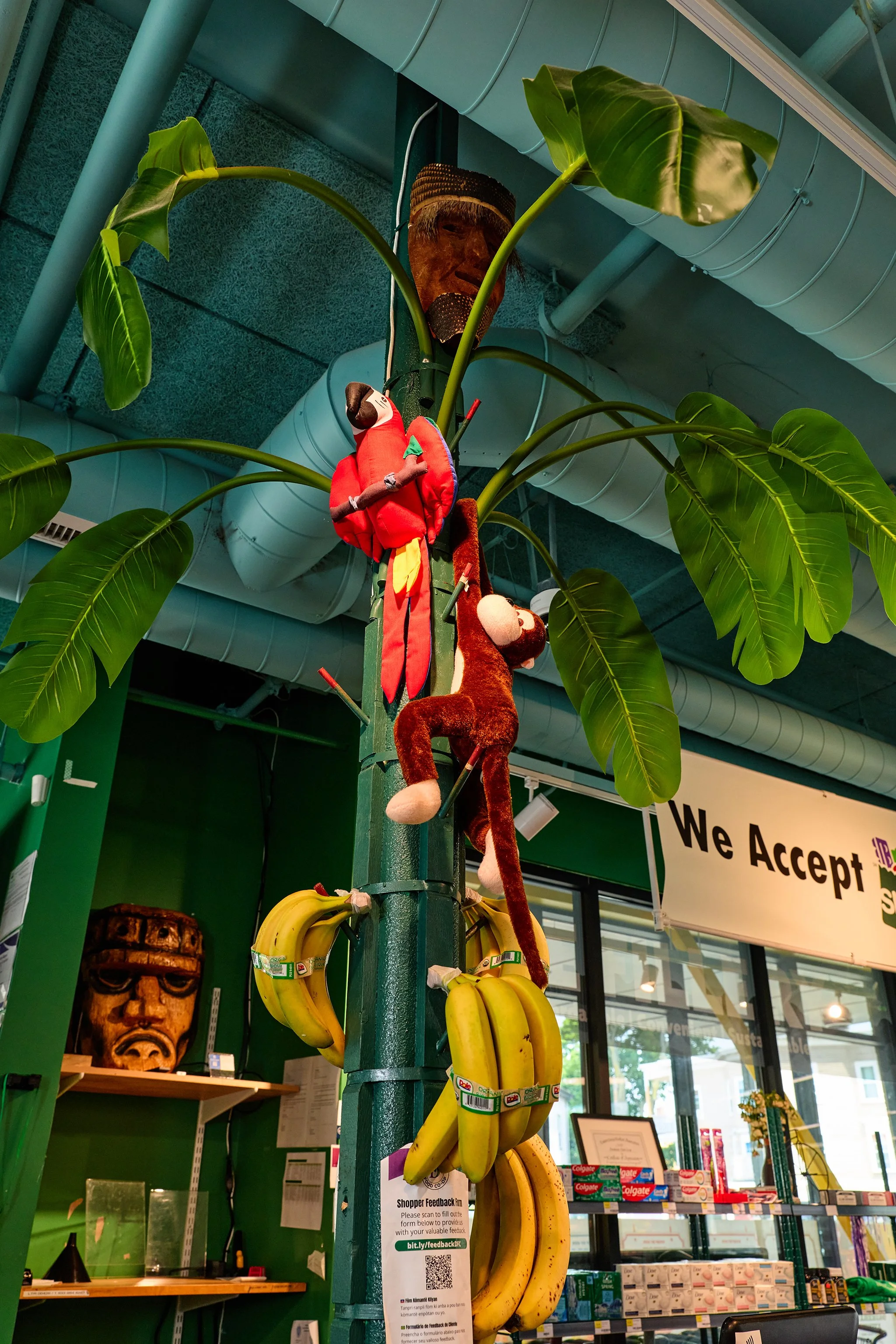 A store display featuring bunches of bananas hanging from a green pole, plush toys attached, and tropical leaves. There are also decorative masks and a sign indicating acceptance of feedback.