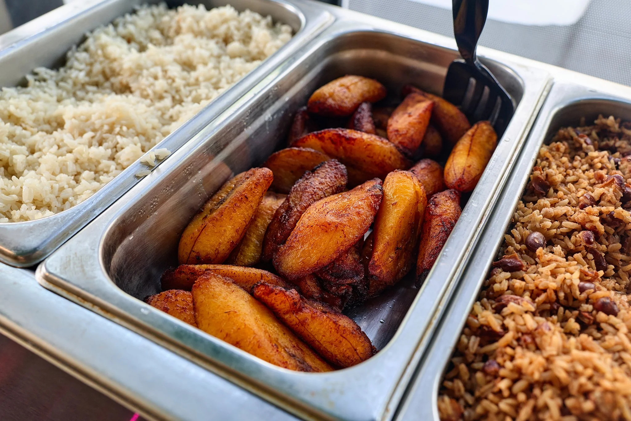 Stainless steel buffet tray containing fried plantains, with other side dishes visible beside it.