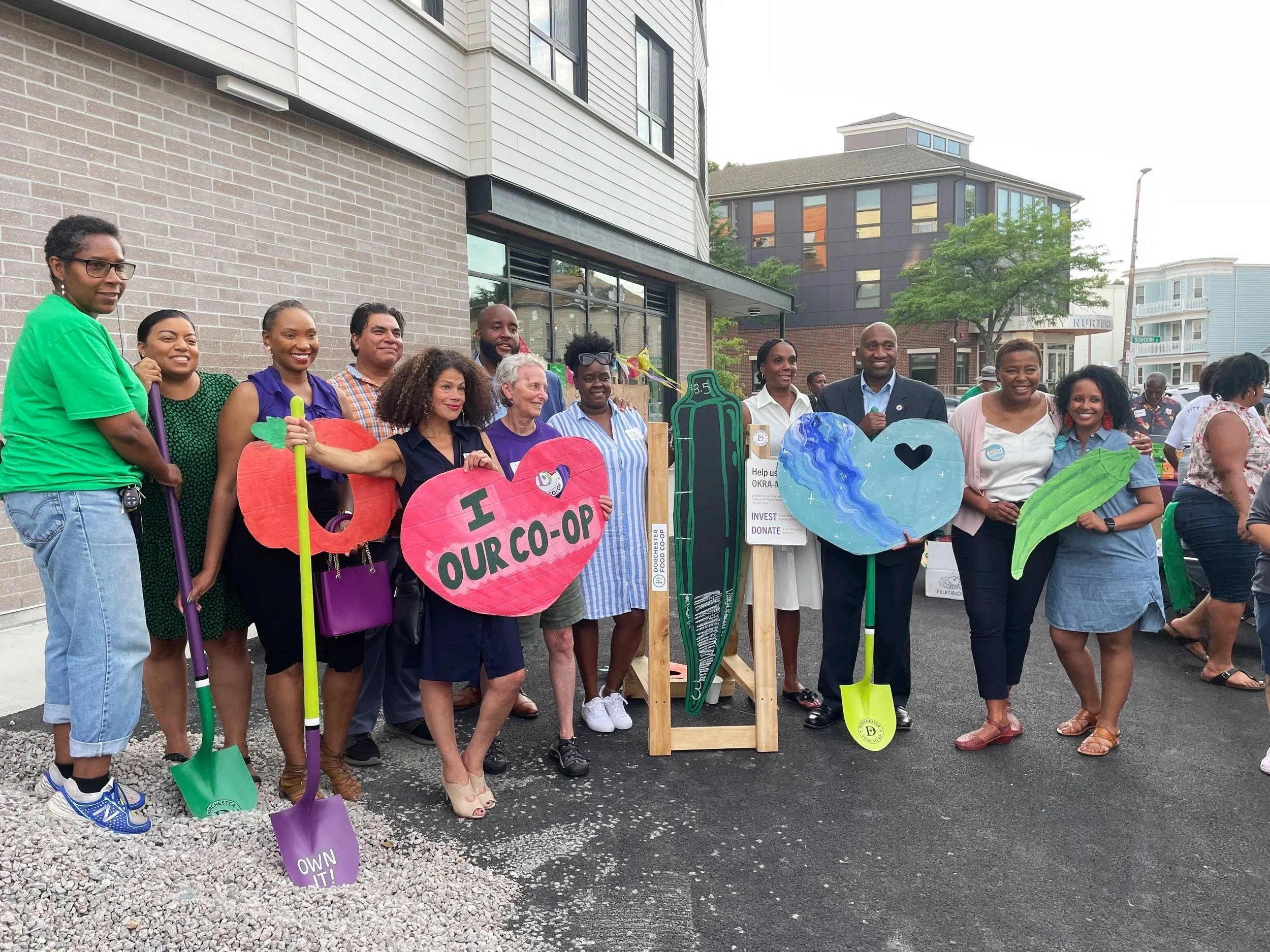 A diverse group of people standing outdoors in front of modern buildings during daytime, holding colorful signs and shovels, participating in a community event or protest.