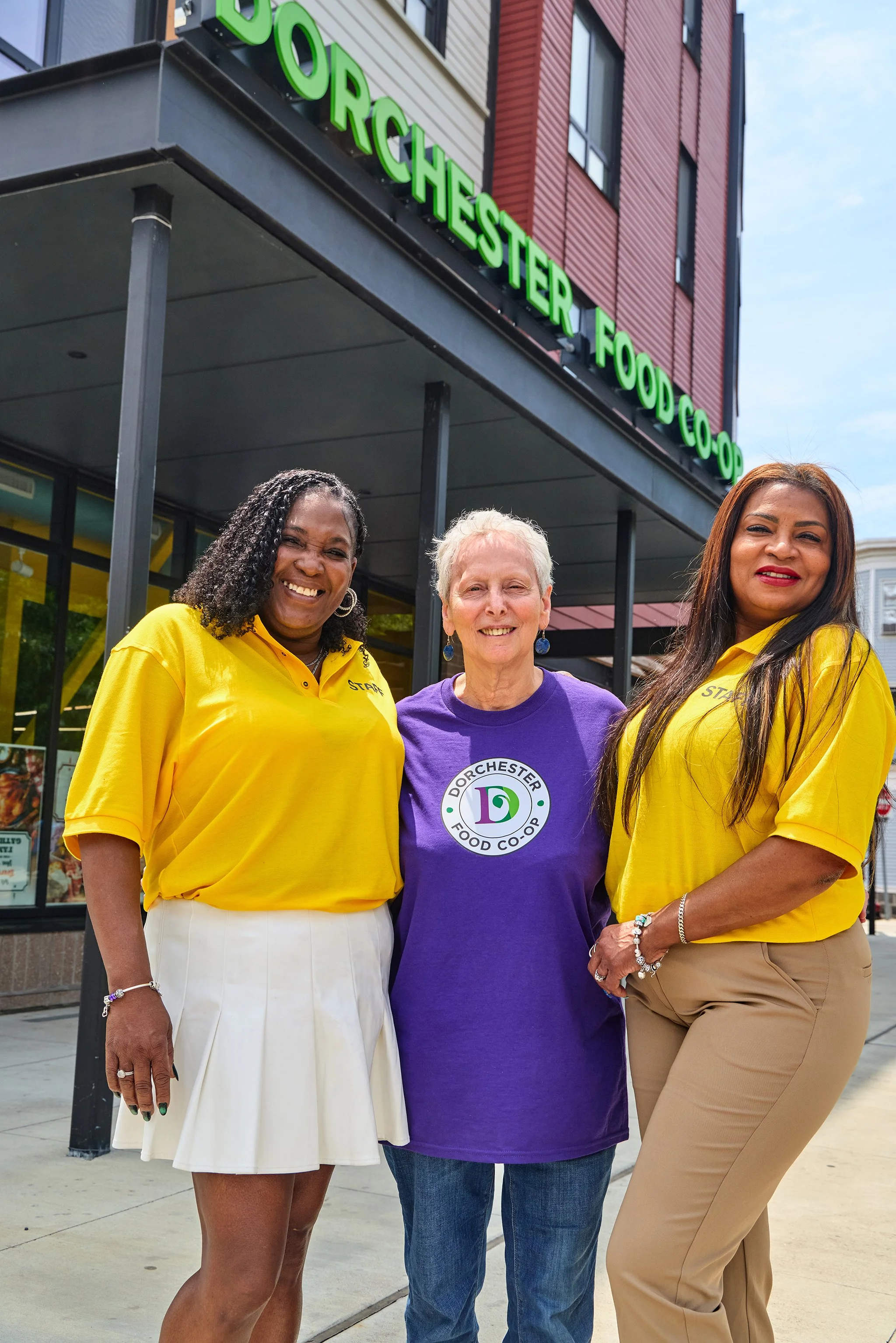 Three women standing in front of a food co-op store, smiling. Two women are wearing yellow shirts with 'STAFF' on them, and one is wearing a purple shirt with a 'Dorchester Food Co-op' logo. The store has a green sign reading 'Dorchester Food Co-op' and large windows.