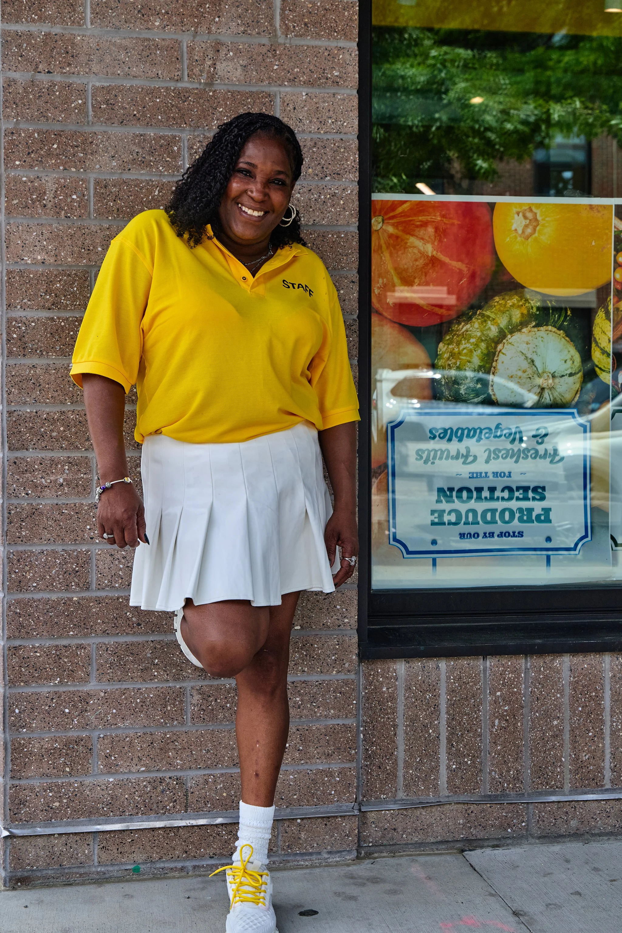 A woman wearing a yellow staff polo shirt, white pleated skirt, and bright yellow sneakers with white socks stands against a brick wall near a window displaying images of various vegetables and fruits.
