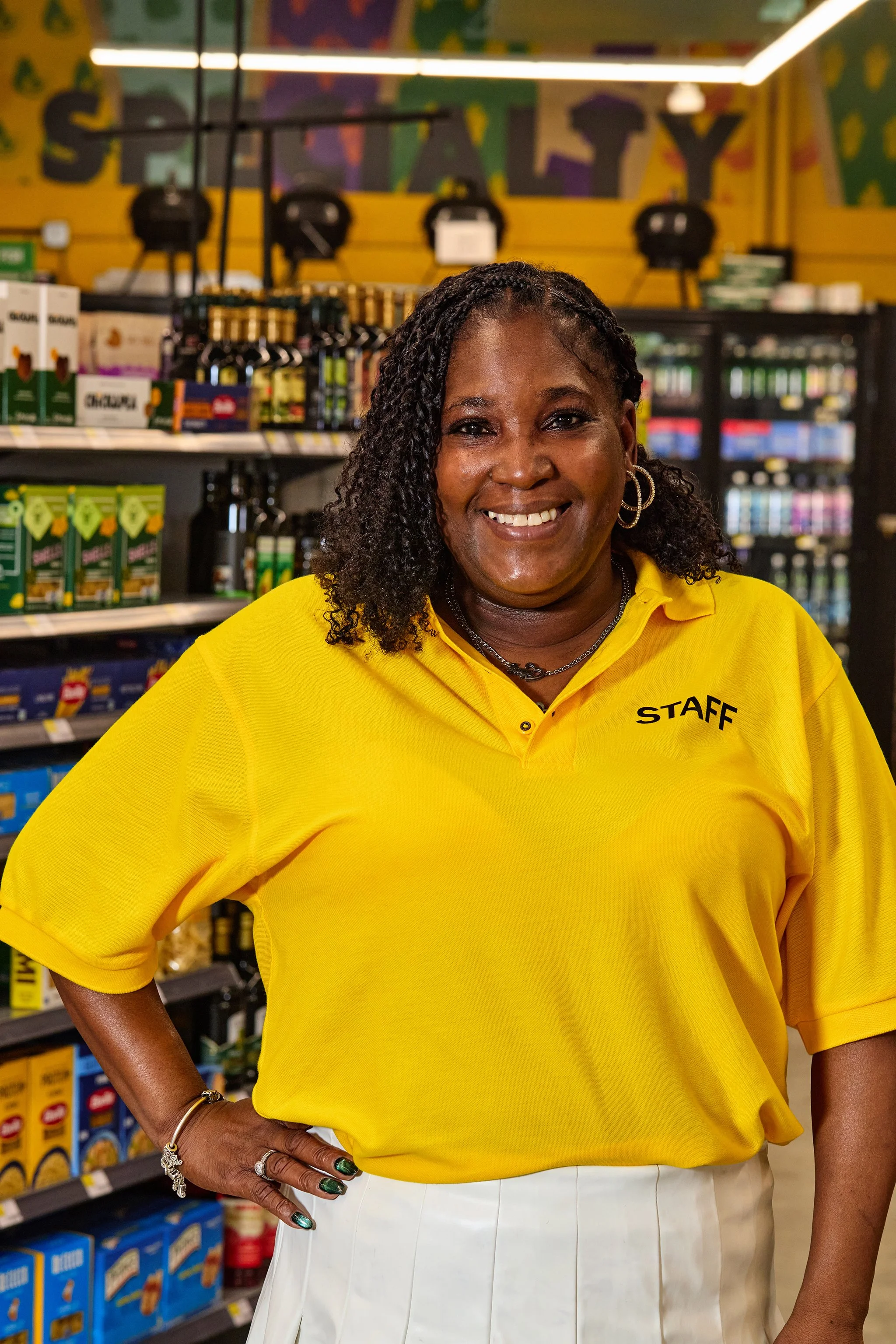 A woman wearing a yellow staff shirt and white skirt poses inside a store aisle, smiling at the camera with her left hand on her hip, accessorized with earrings, rings, and a bracelet.