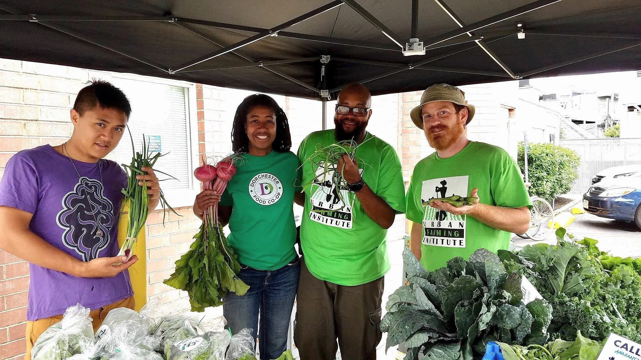 Four people standing together at a farmers market stall with fresh vegetables, smiling, with a brick building and parked cars in the background.
