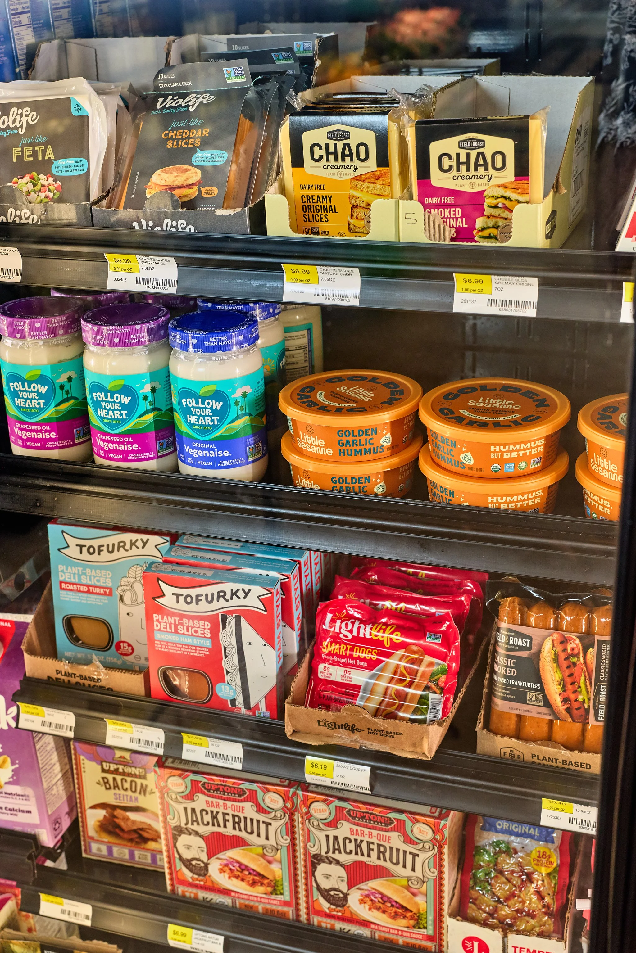 Shelf display with plant-based deli slices, cheese slices, hummus, hot dog buns, and frozen dinner boxes.