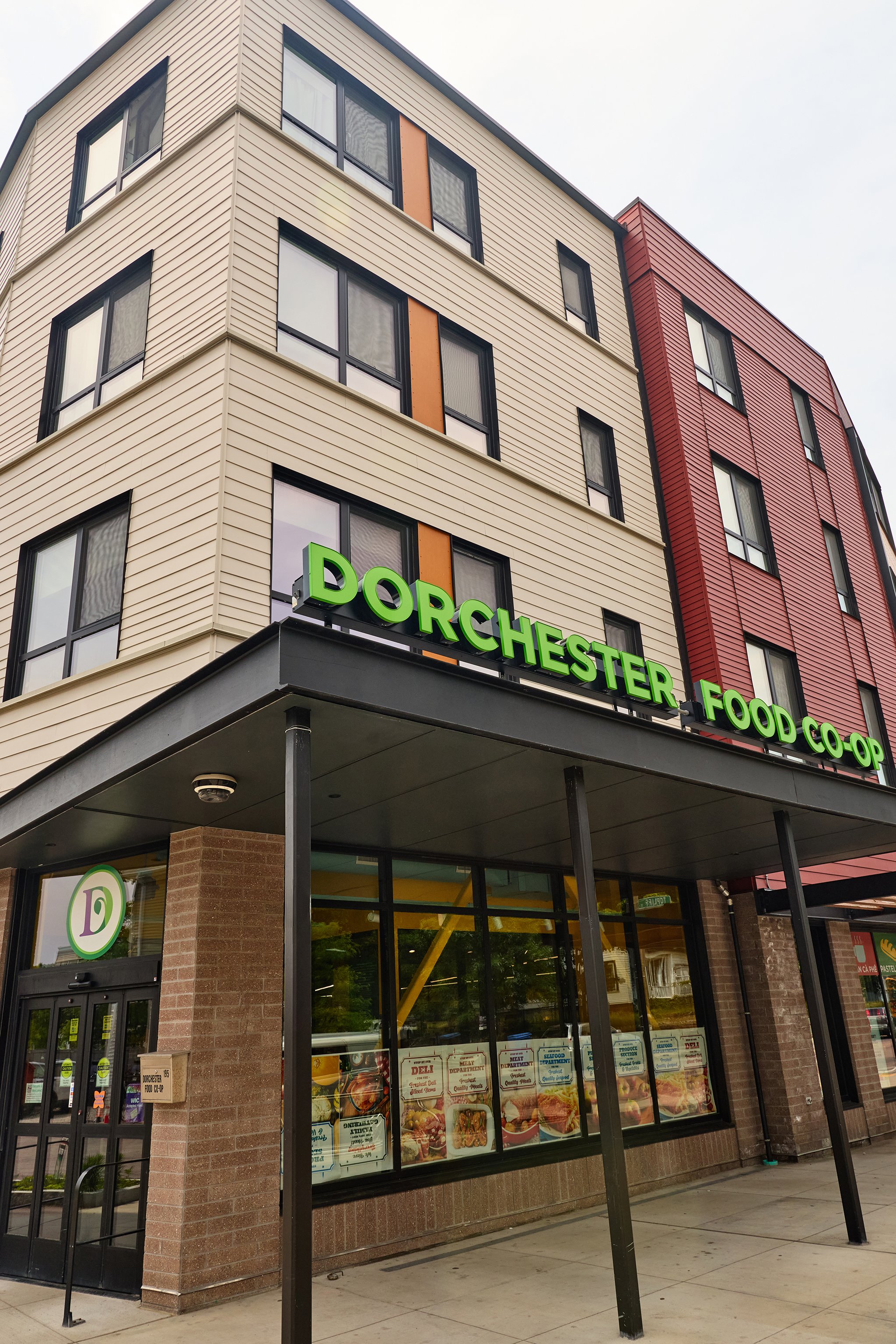 Exterior of a grocery store named Dorchester Food Co-op, with large windows displaying food posters and a glass door entrance. The building has beige and red siding, with black metal accents and a sign in green letters.