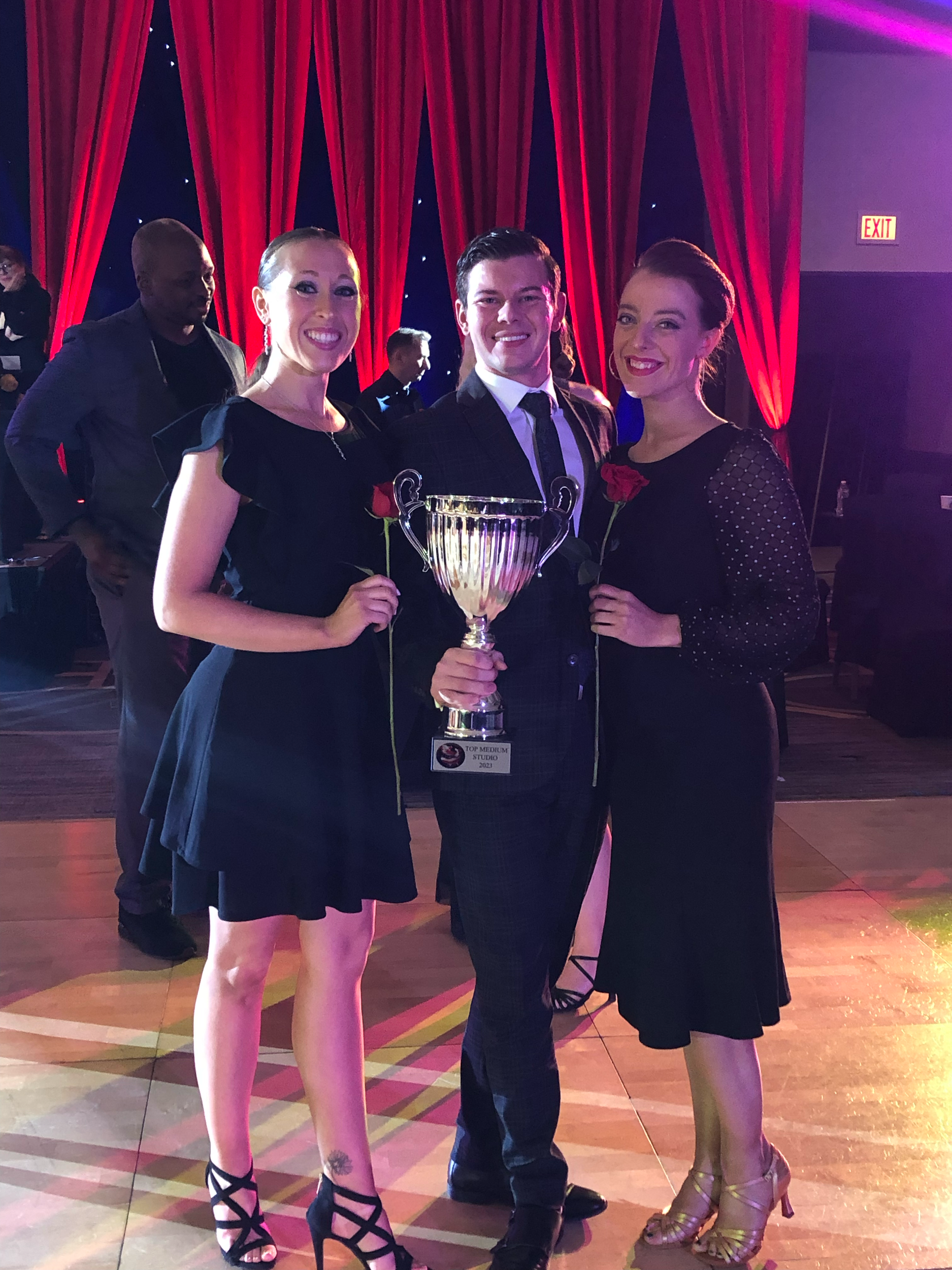 Three women at an award ceremony, one holding a large trophy, standing under red curtains with dim lighting.