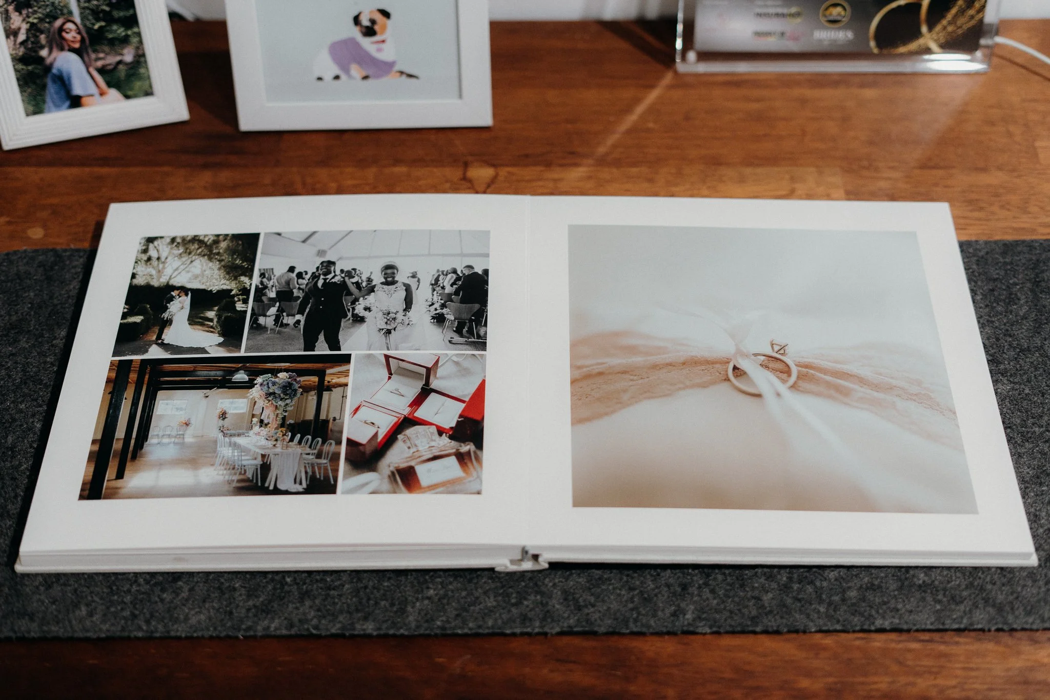 Open wedding photo album on a wooden table displaying photos of a wedding, including a bride and groom walking, a ceremony, a decorated table, and jewelry on a ring pillow.