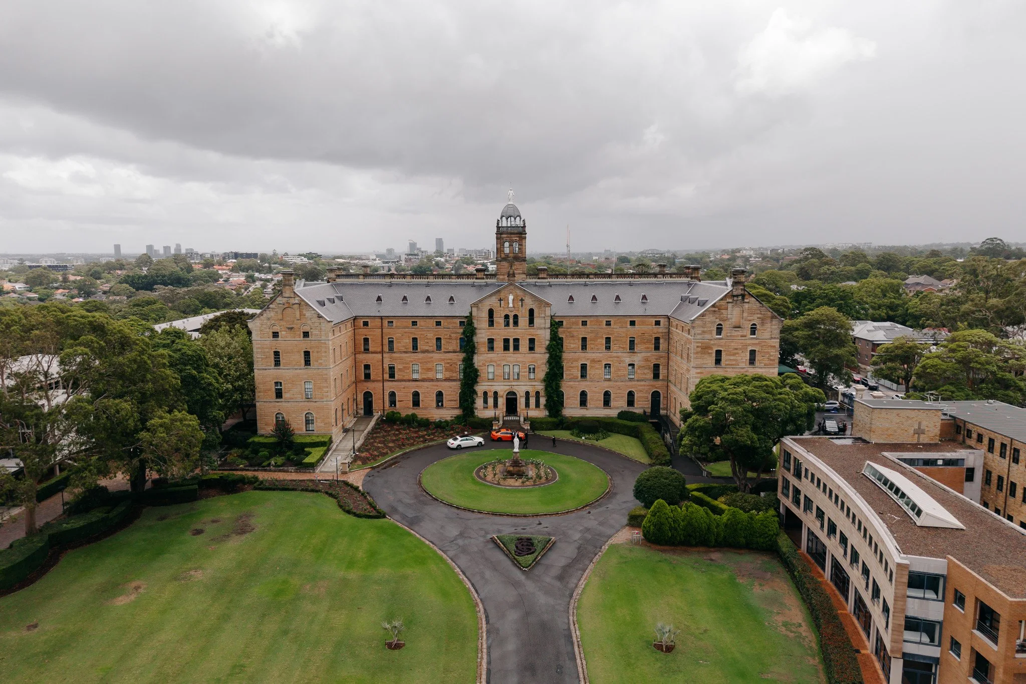 A historic brick building with a central tower, surrounded by trees and a circular driveway with parked cars, on an overcast day.