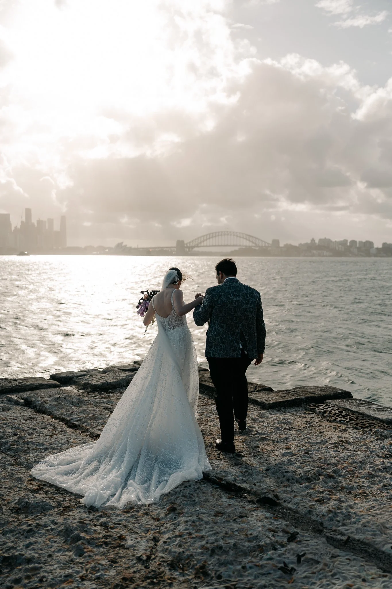 A bride and groom walking by the water during sunset, with the city skyline and bridge in the background.