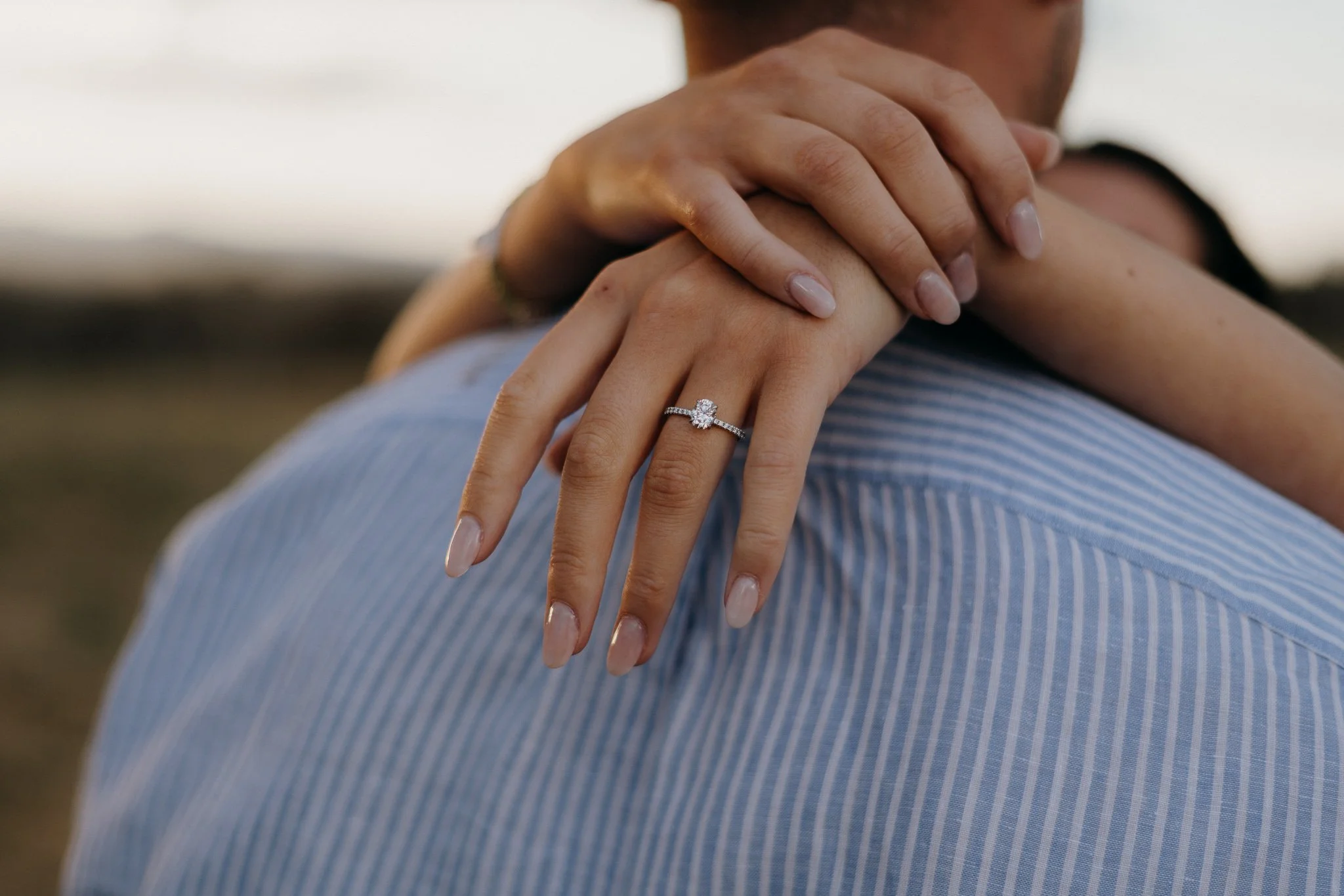 A woman's hand with an engagement ring resting on a man's shoulder, with both of them embracing. The focus is on the ring and hands, with a blurred outdoor background.