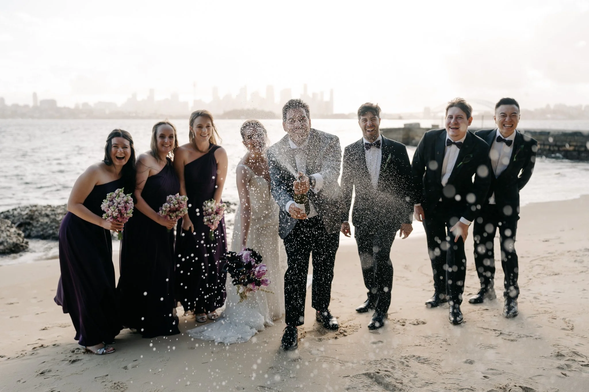 A group of bridesmaids and groomsmen celebrating their wedding on a beach, spraying champagne, with city skyline in the background.