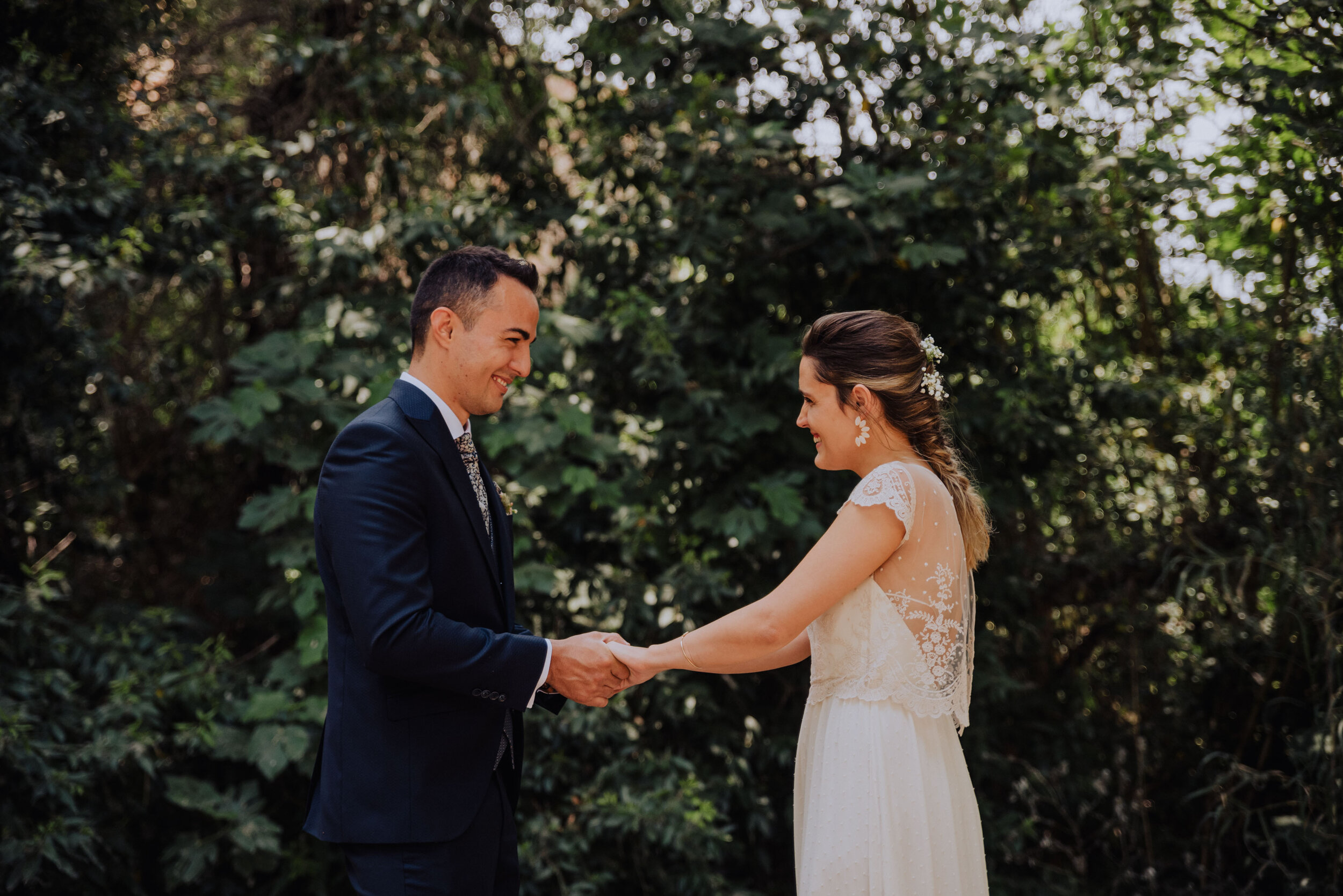 Pareja de novios tomados de la mano en un jardín, él en traje oscuro, ella en vestido blanco de encaje. Fotógrafo de bodas en Sant Celoni y Barcelona.