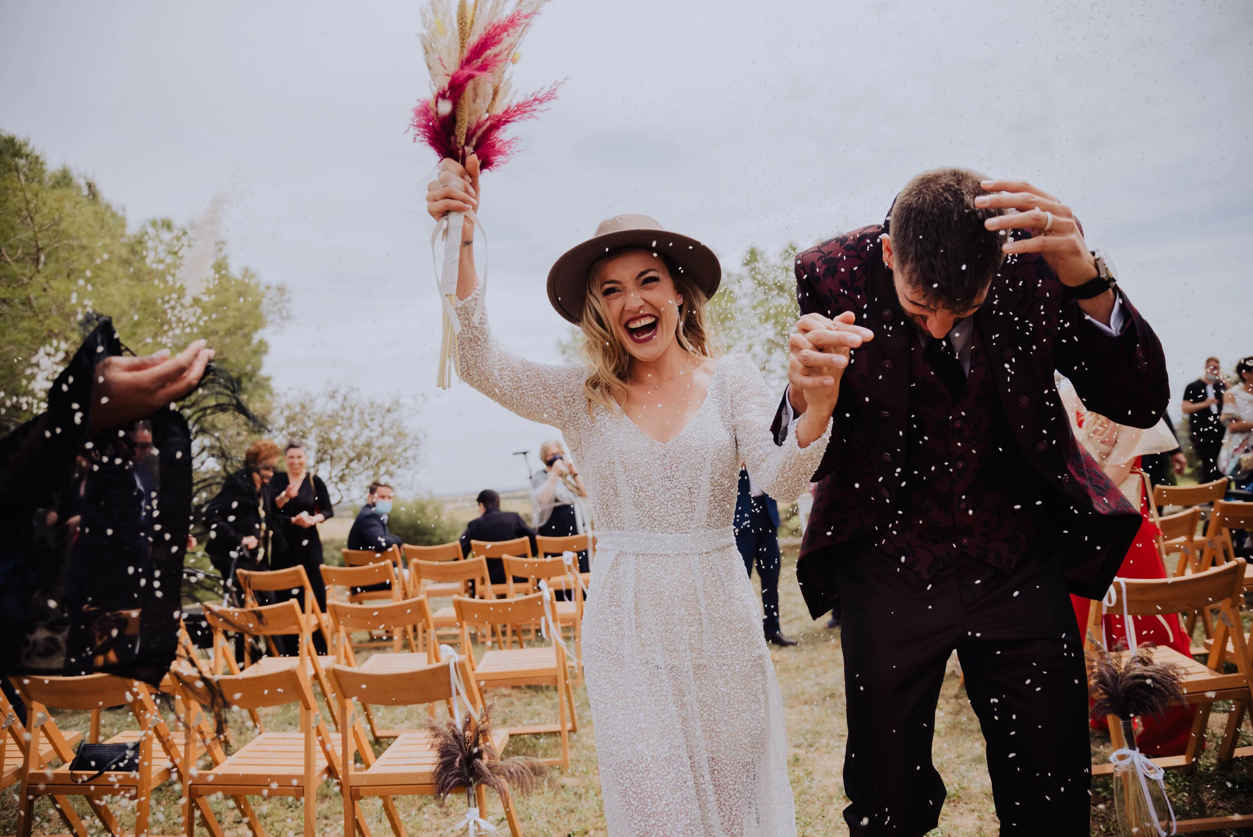 Pareja recién casada caminando por el pasillo mientras les lanzan confeti, con sillas y personas al fondo. Fotógrafo de bodas en Sant Celoni (Barcelona y Catalunya)