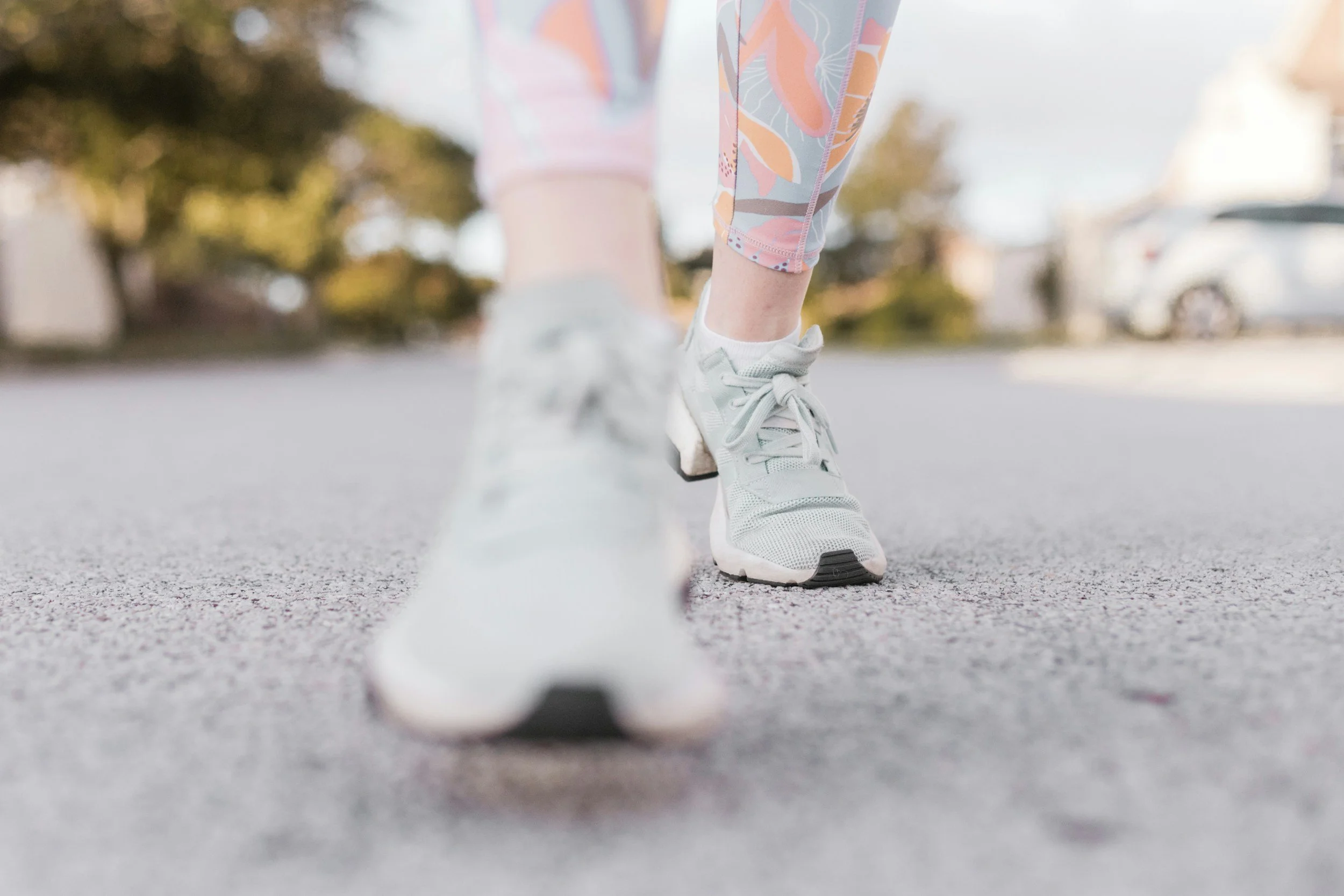 Close-up of a person wearing light gray running shoes and colorful leggings, preparing to run on an outdoor street with trees and parked cars in the background.