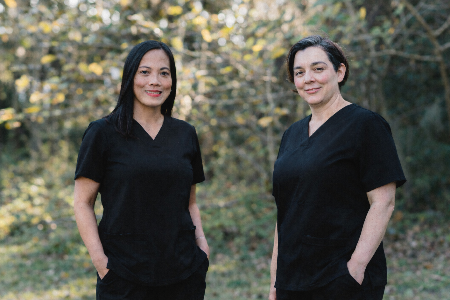 Marj (PT) on the left and Lisa (OT) on the right, both wearing black scrubs with a wooden background