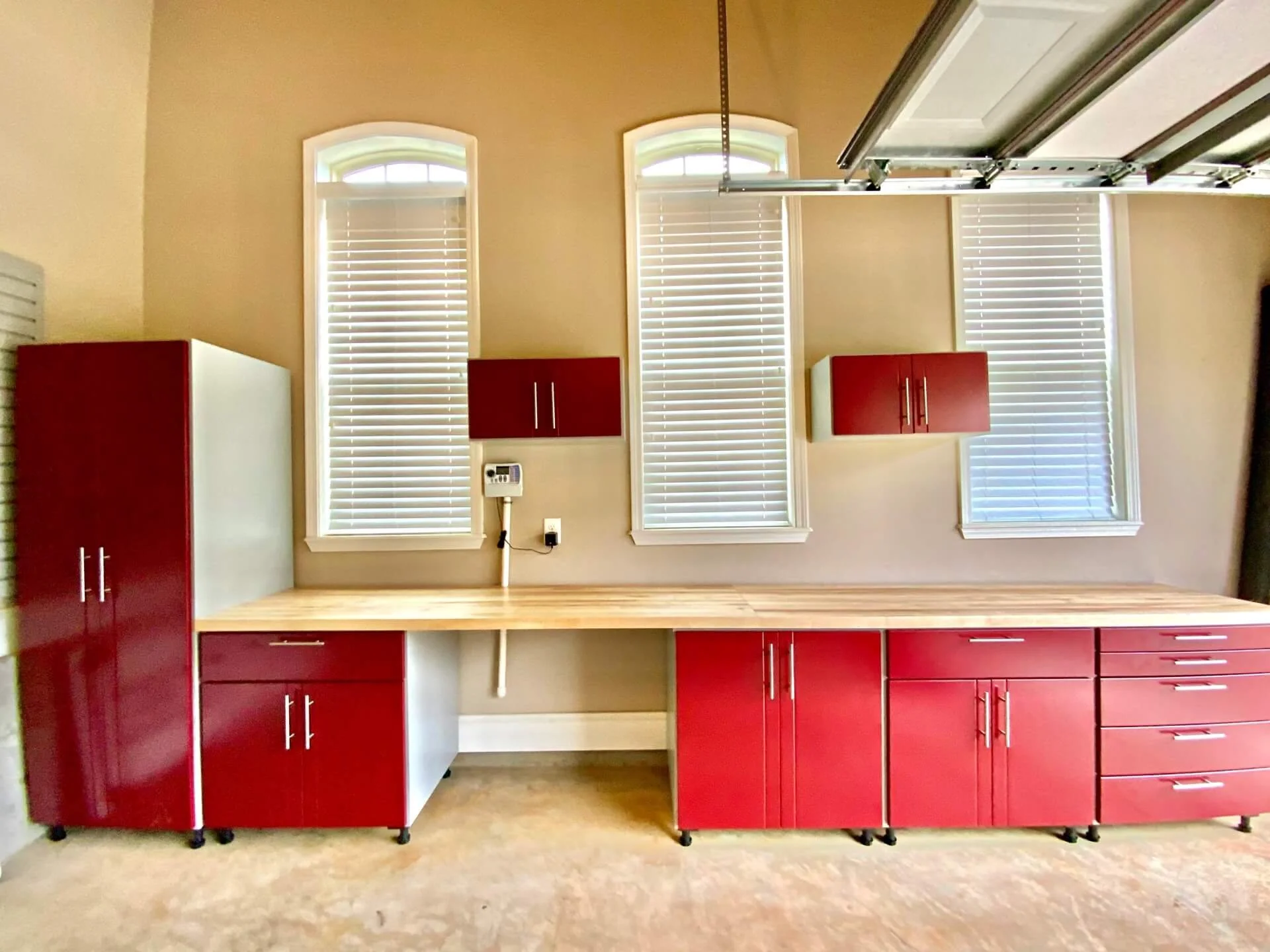 Empty kitchen with red and white cabinets, three tall windows with blinds, beige walls, and a wooden countertop.