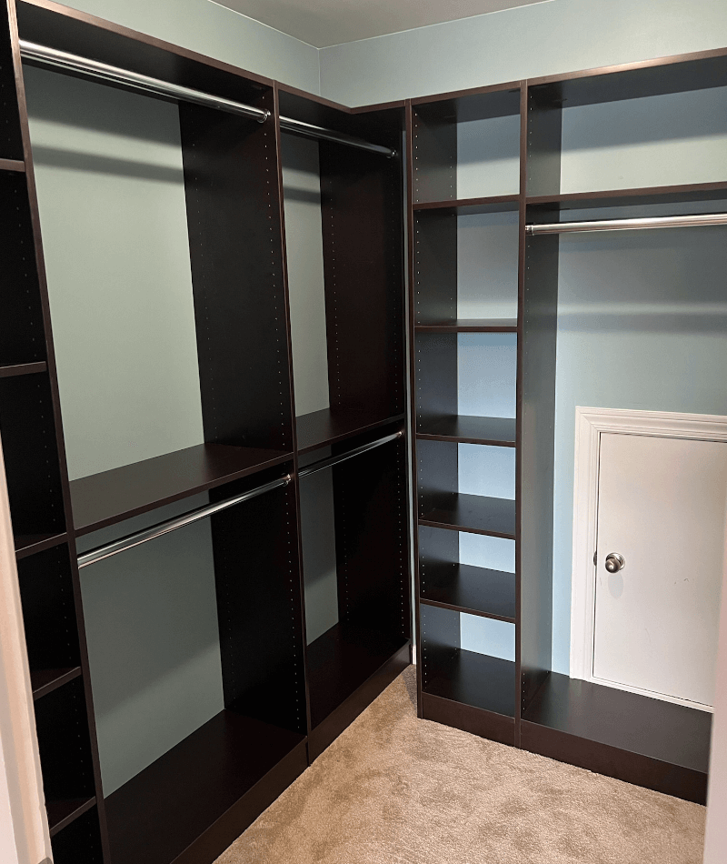 Empty walk-in closet with black shelving and hanging rods, beige carpet, light blue walls, small white access door.