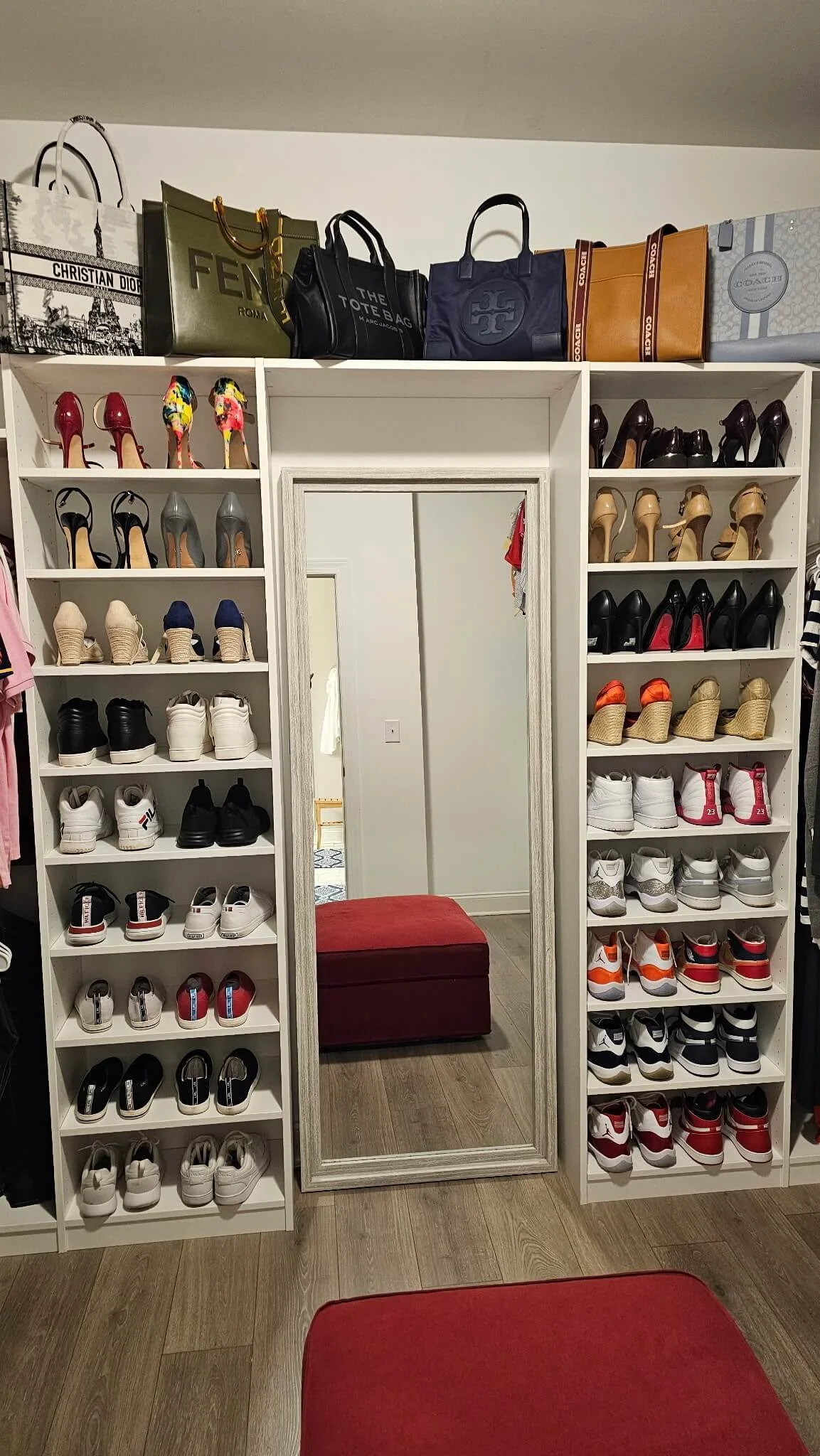 A shoe and handbag closet with various shoes organized on shelves and handbags on top, a full-length mirror, and a red ottoman in front.