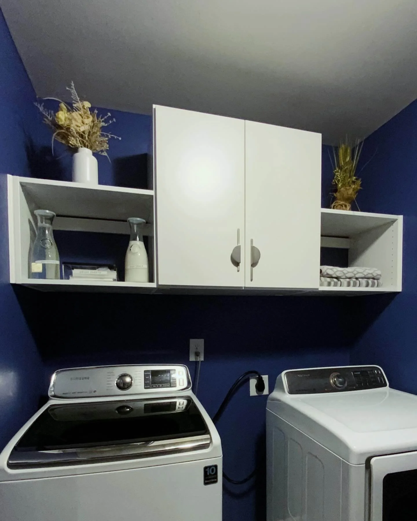 White laundry washer and dryer placed side by side in a laundry room with dark blue walls. A white shelf mounted above holds a white vase with dried plants on either side.