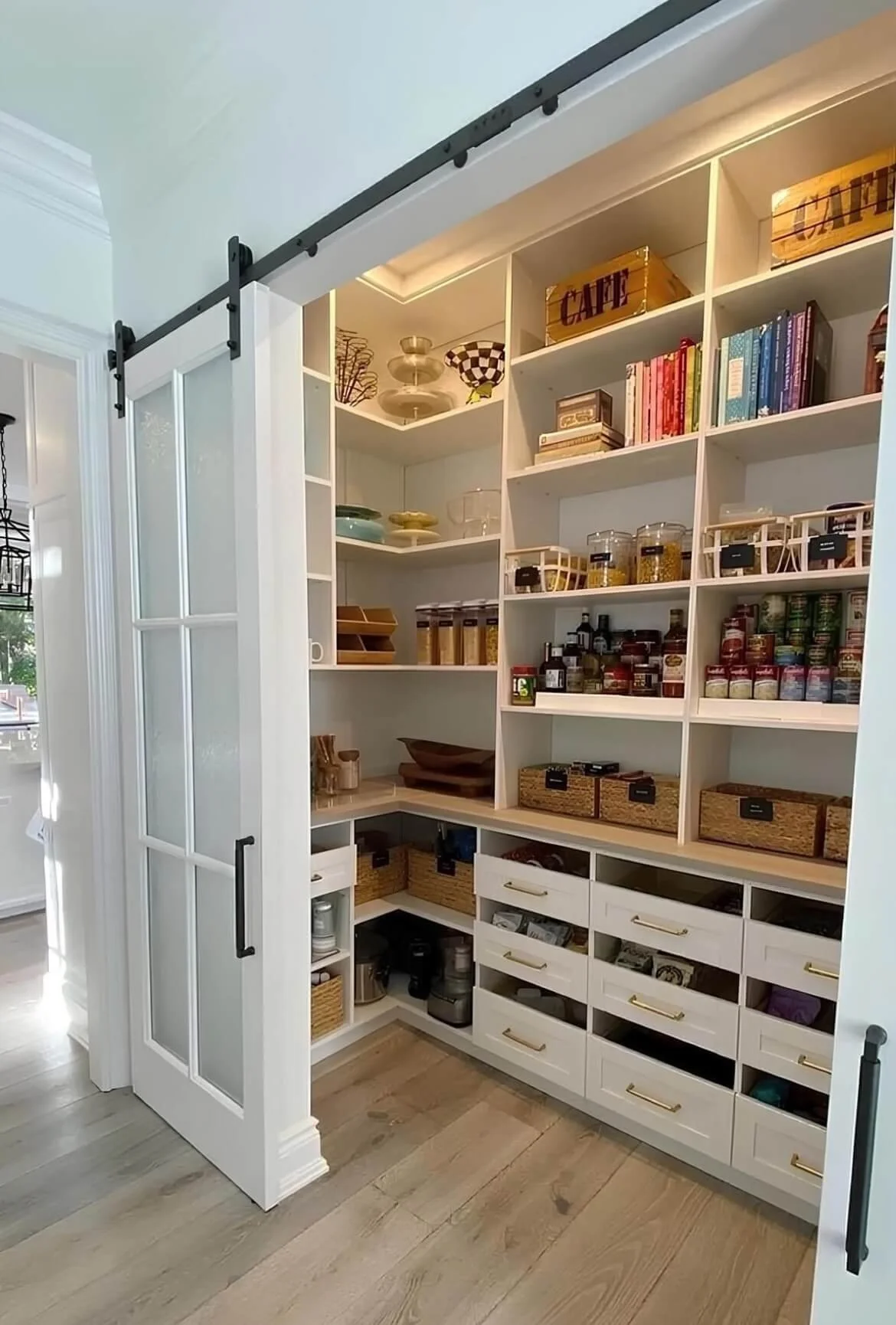 View of a pantry with white shelves filled with cooking and baking ingredients, utensils, and cookbooks, enclosed by sliding barn door on a metal track.