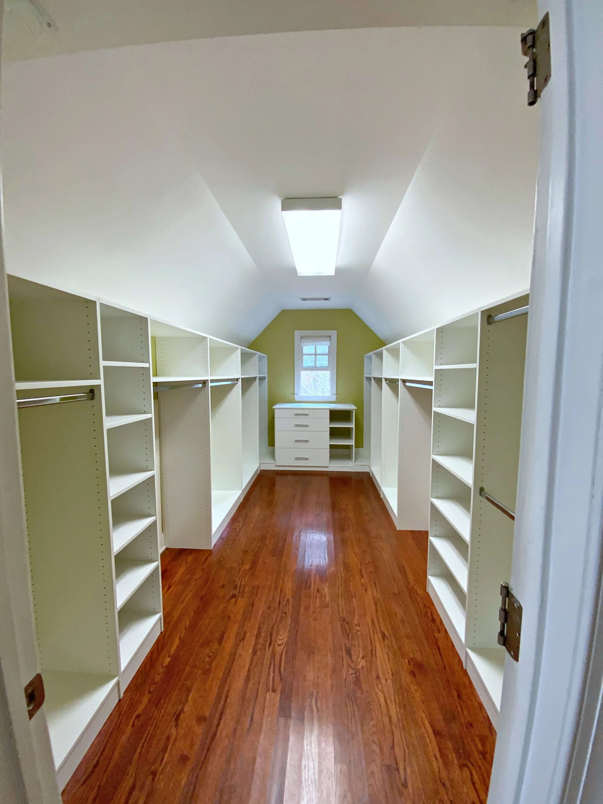 A walk-in closet with white shelves on both sides, a small window at the back, and a wooden floor with a medium finish.