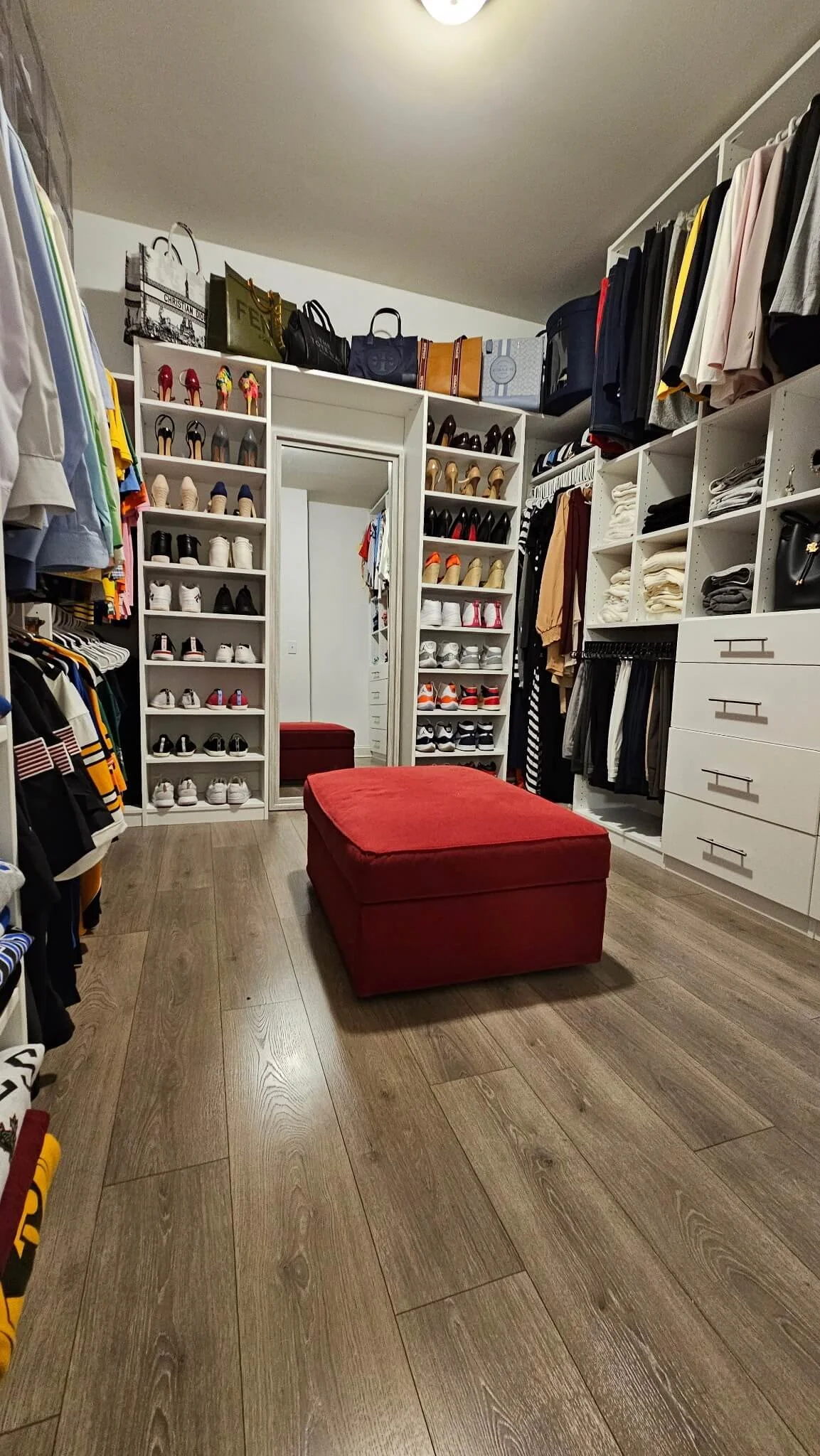 Walk-in closet with organized shelves of shoes, folded clothes, handbags, and hanging garments. There is a red ottoman in the center and a mirror on the back wall.