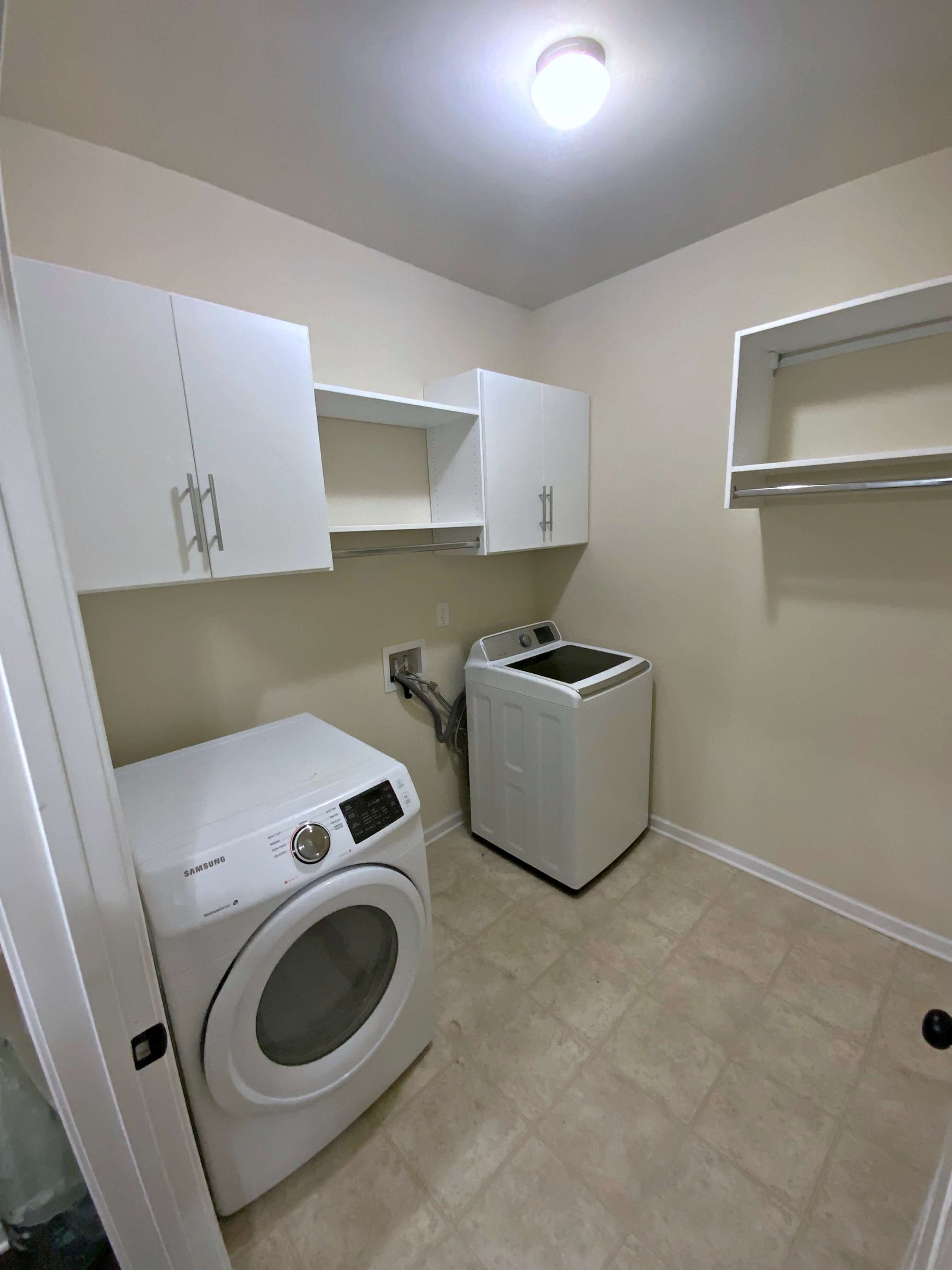 A laundry room with a front-loading Samsung washing machine, a compact top-loading dryer, white wall-mounted cabinets, a folding shelf, and a metal rod for hanging clothes. The room has beige walls, a ceiling light, and tiled flooring.