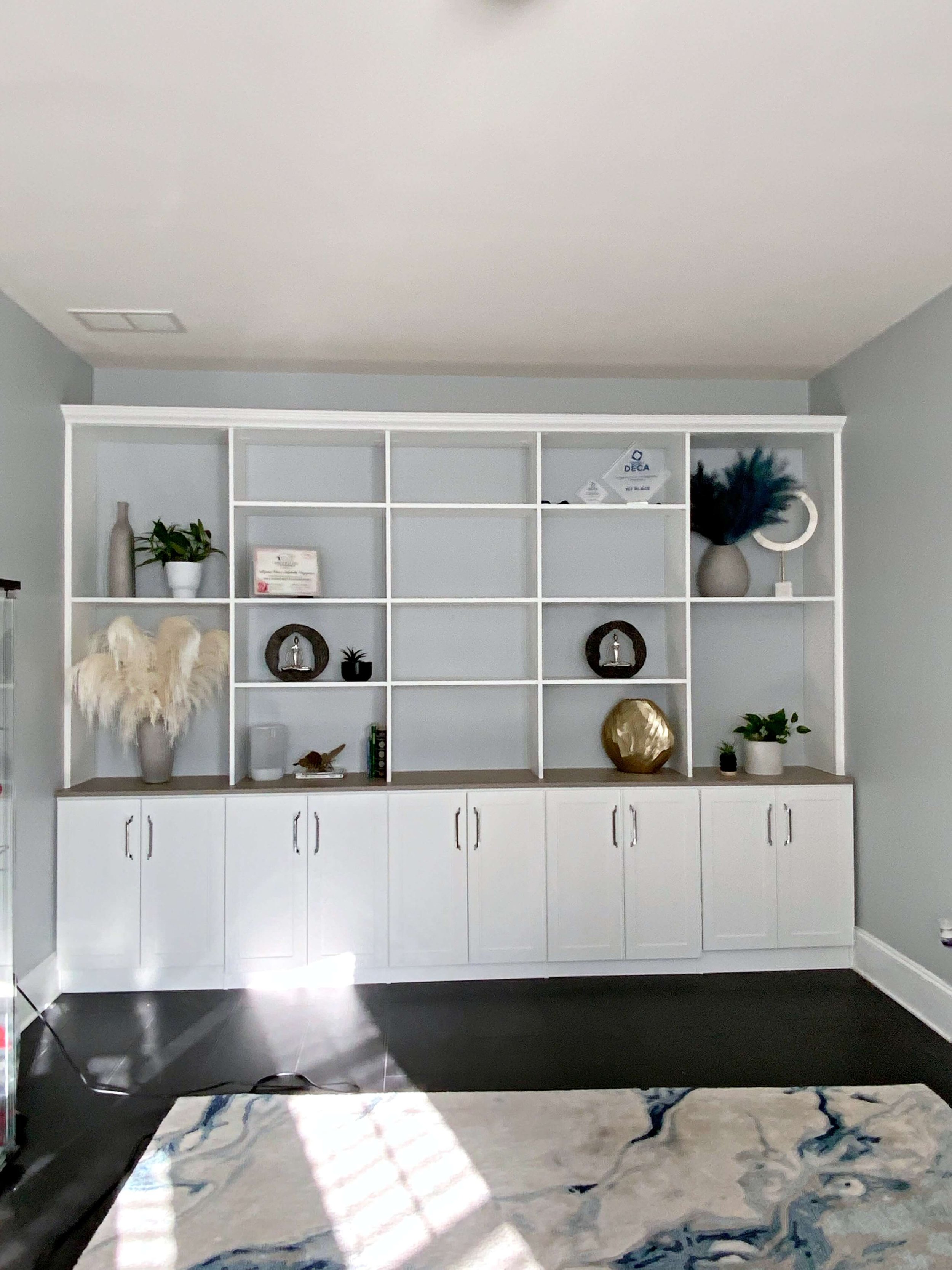 A white bookshelf with decorative items such as plants, books, framed pictures, and sculptures, located against a light gray wall. There is a large potted pampas grass on the left side of the shelf, and the lower part of the bookshelf has closed cabinets. The floor appears to be dark hardwood or laminate, and there is a marble or stone rug with a white and blue pattern in front of it.