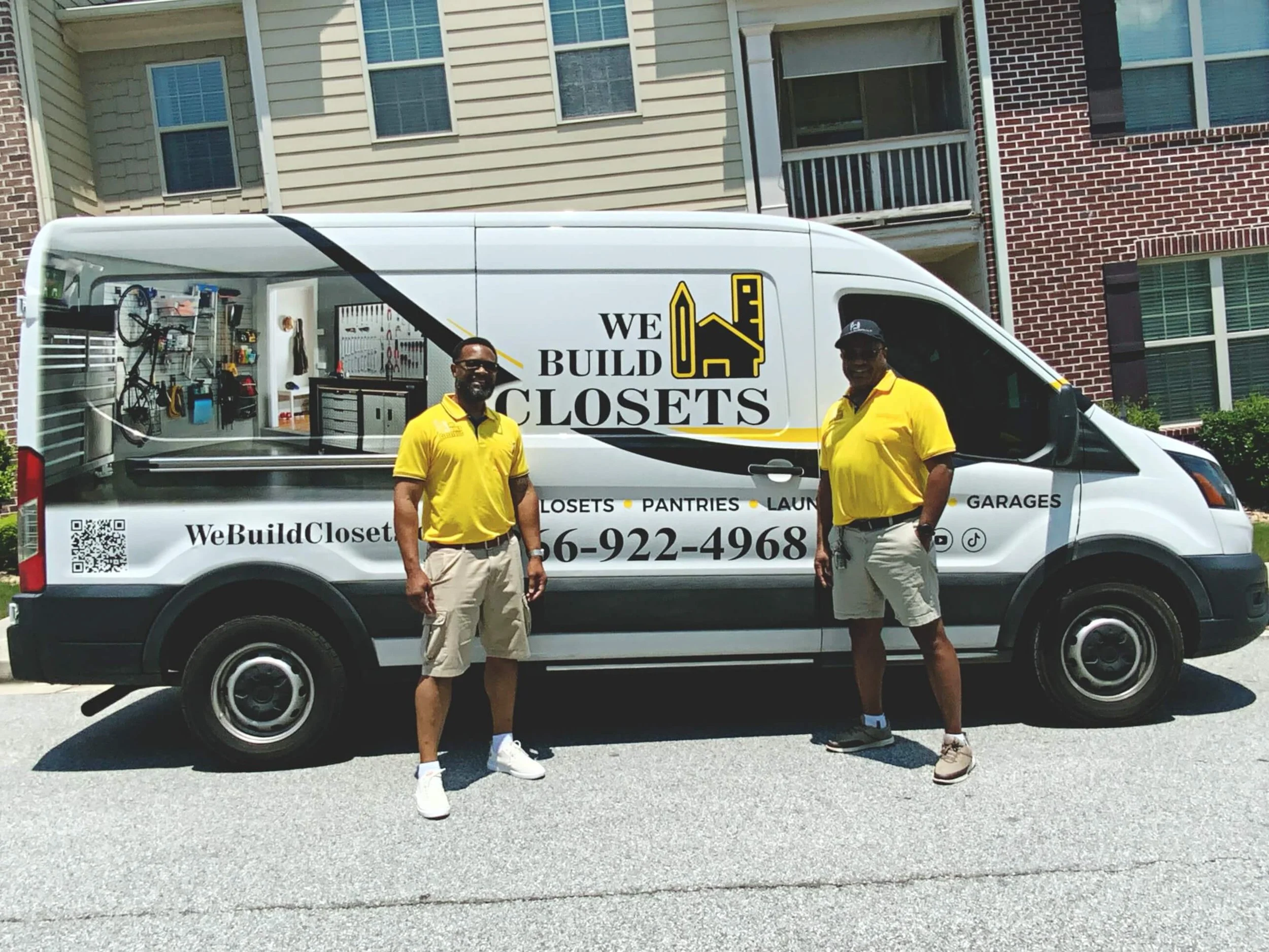 Two men in yellow polo shirts standing in front of a cargo van with an open side door revealing tools and storage shelves. The van has the logo 'We Build Closets' and a phone number displayed on the side.