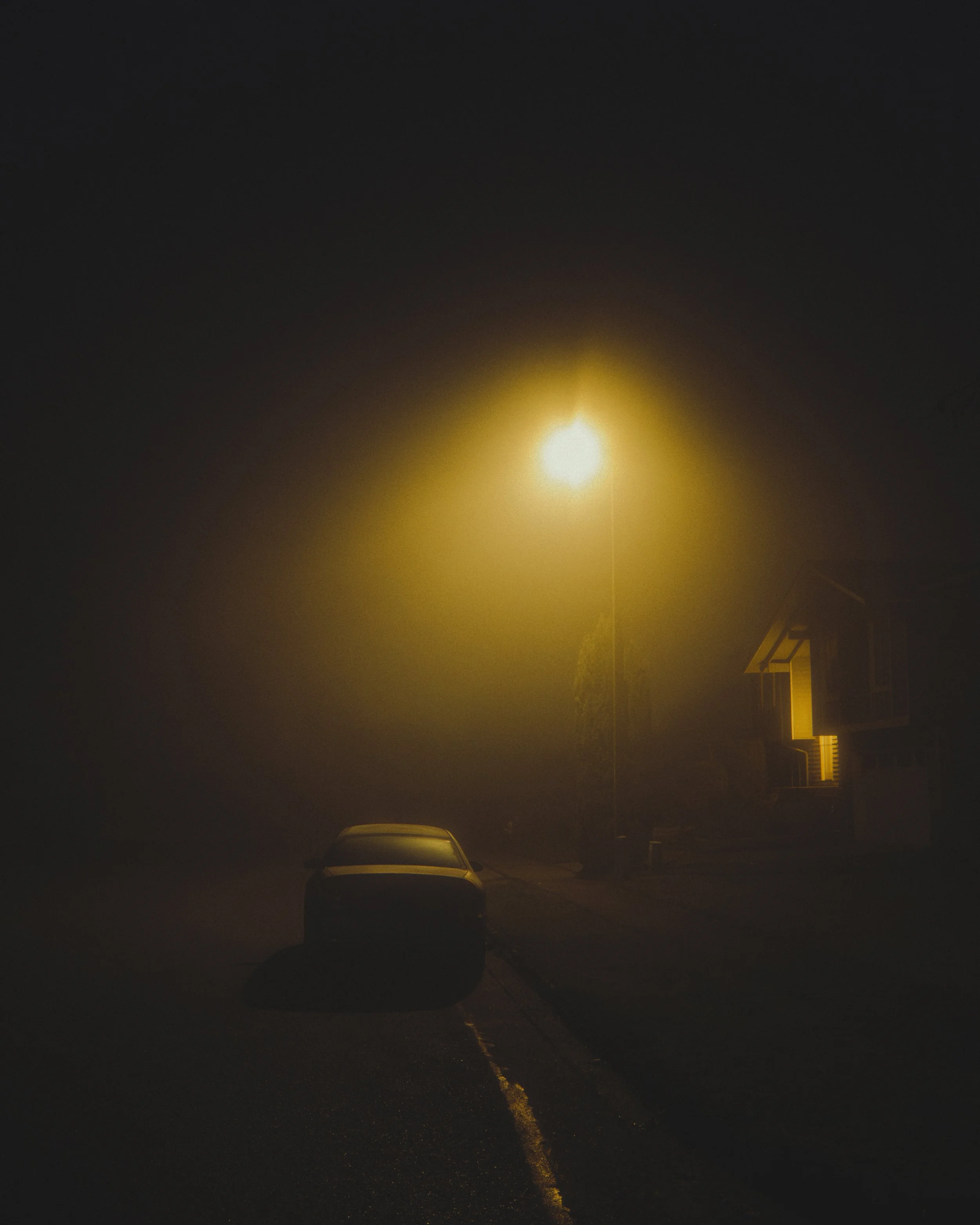 A dark street at night illuminated by a single yellow streetlamp with a parked car nearby.