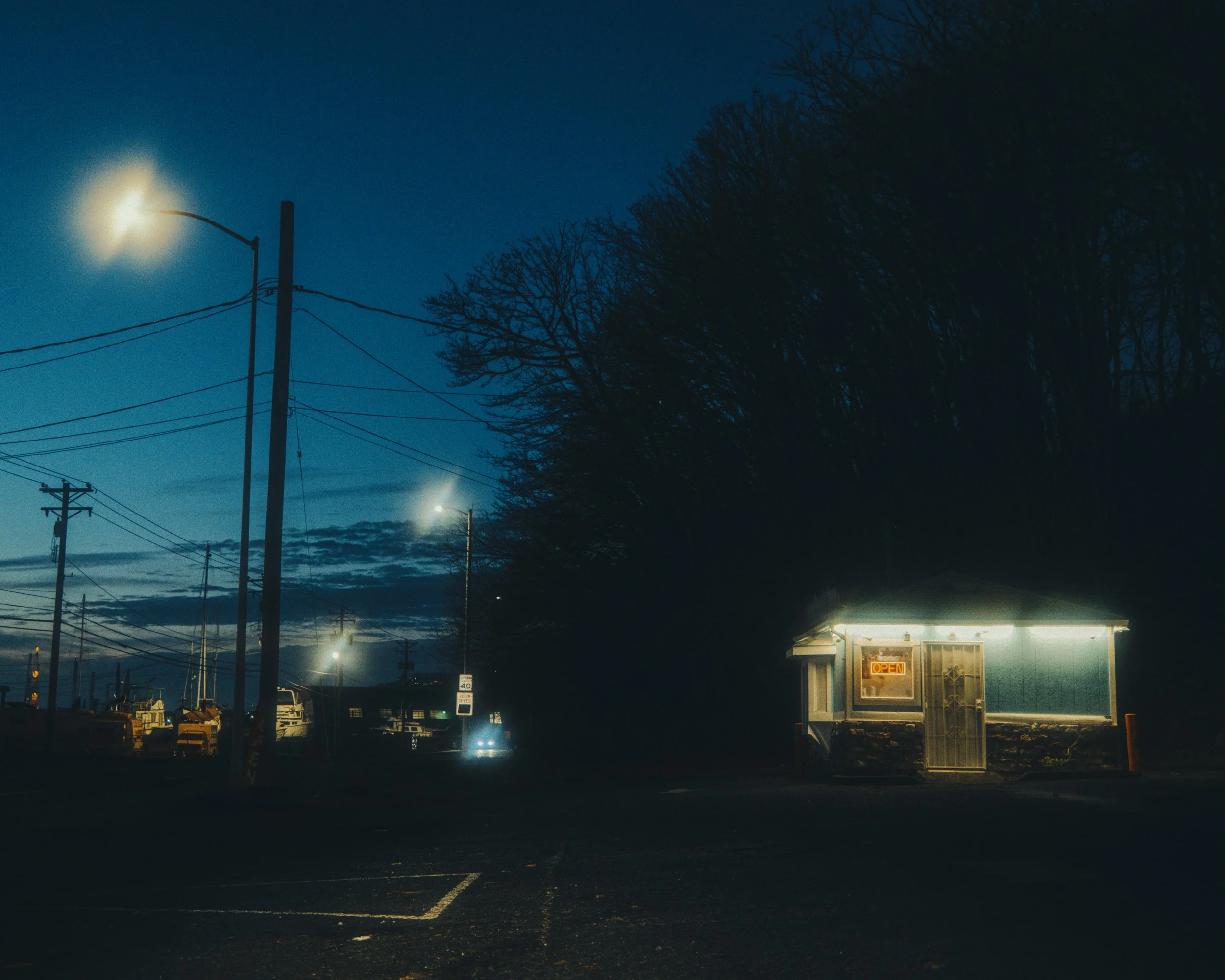 Small, lit-up building with an 'OPEN' sign in a dark parking lot at dusk, with trees and power lines in the background.