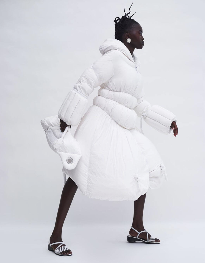 A woman dressed in a white puffy winter coat with oversized sleeves and a full skirt, wearing white sandals and large pearl earrings, walking in profile against a plain white background.