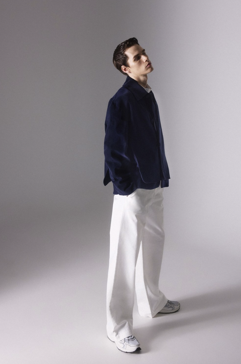 A young man with dark hair styled back, wearing a navy blue jacket, white pants, and sneakers, standing against a gray gradient background, looking slightly upward and to the side with a neutral expression.