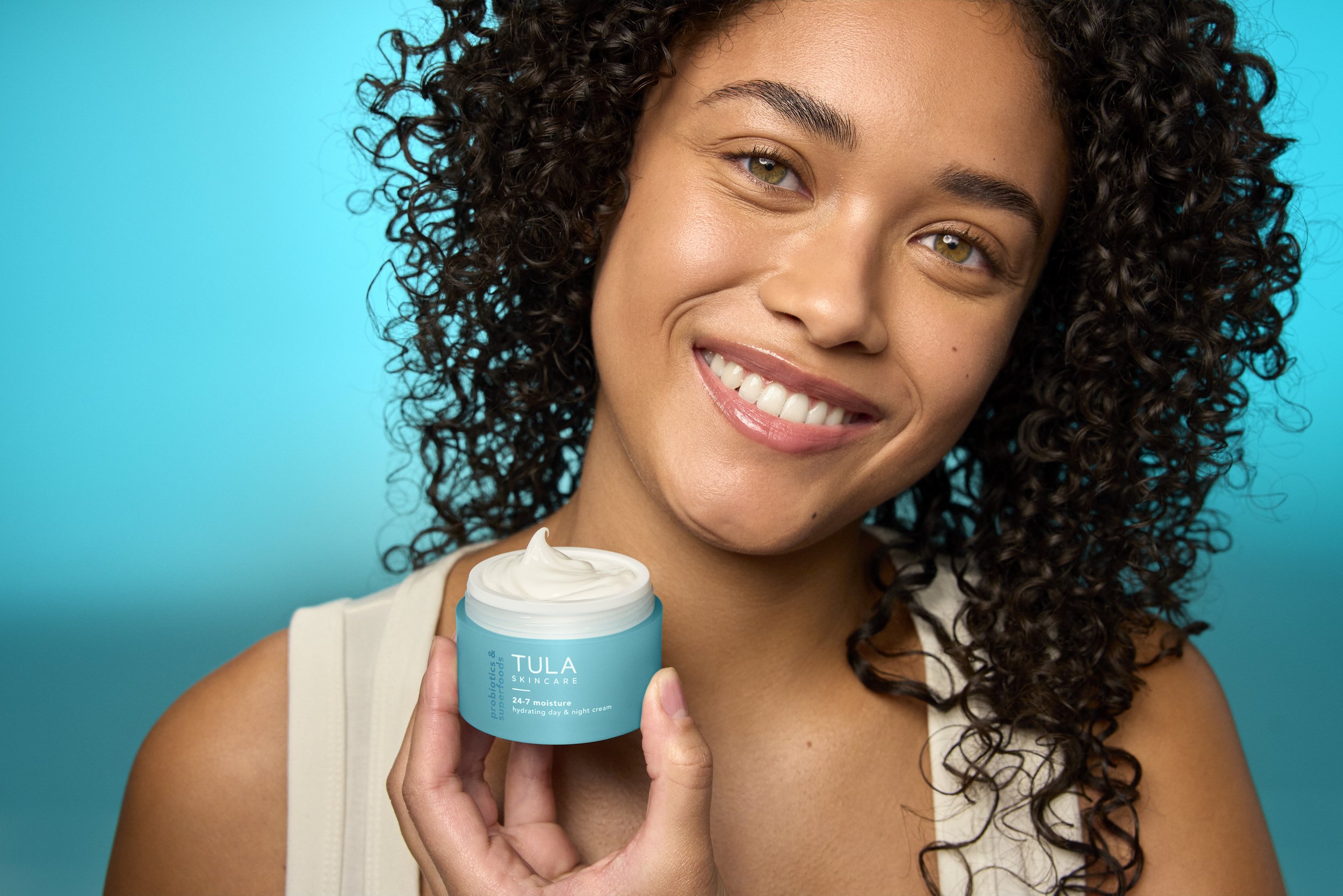 Smiling woman with curly hair holding a blue jar of TULA skincare product against a blue background.