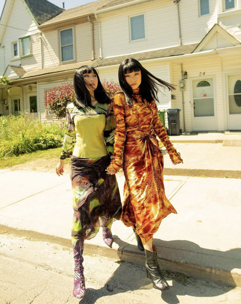 Two fashionable women with black hair and similar hairstyles walk together on a sunny sidewalk in front of a light-colored multi-story house with a garden.