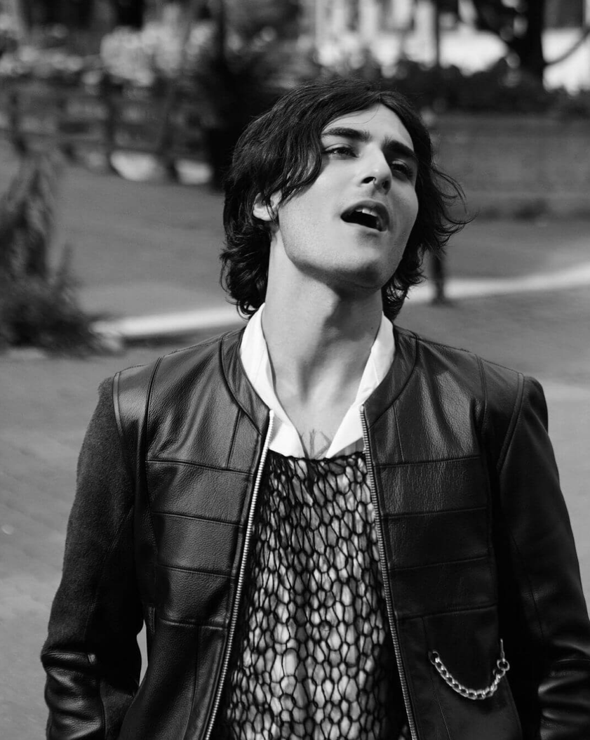 Black and white photo of a young man with shoulder-length hair, wearing a leather jacket over a layered shirt, outdoors with trees and a sidewalk in the background.