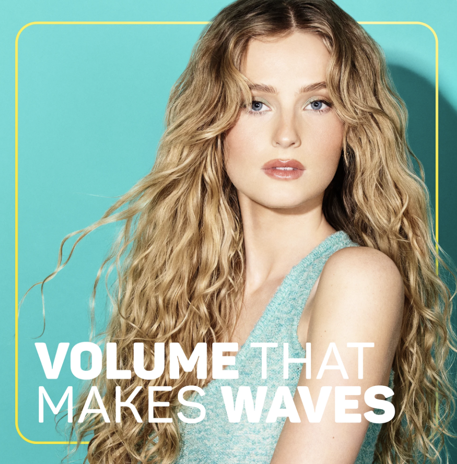 A woman with long, curly blonde hair and blue eyes posing against a bright teal background. She is wearing a sleeveless, light blue top. The text on the image reads 'VOLUME THAT MAKES WAVES' in white and light blue letters.