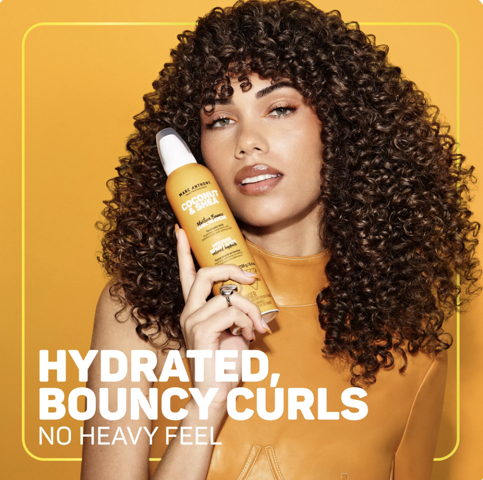 A woman with curly hair holding a bottle of Marc Anthony coconut and shea leave-in conditioner. The background is yellow, and text on the image says 'Hydrated Bouncy Curls No Heavy Feel.'