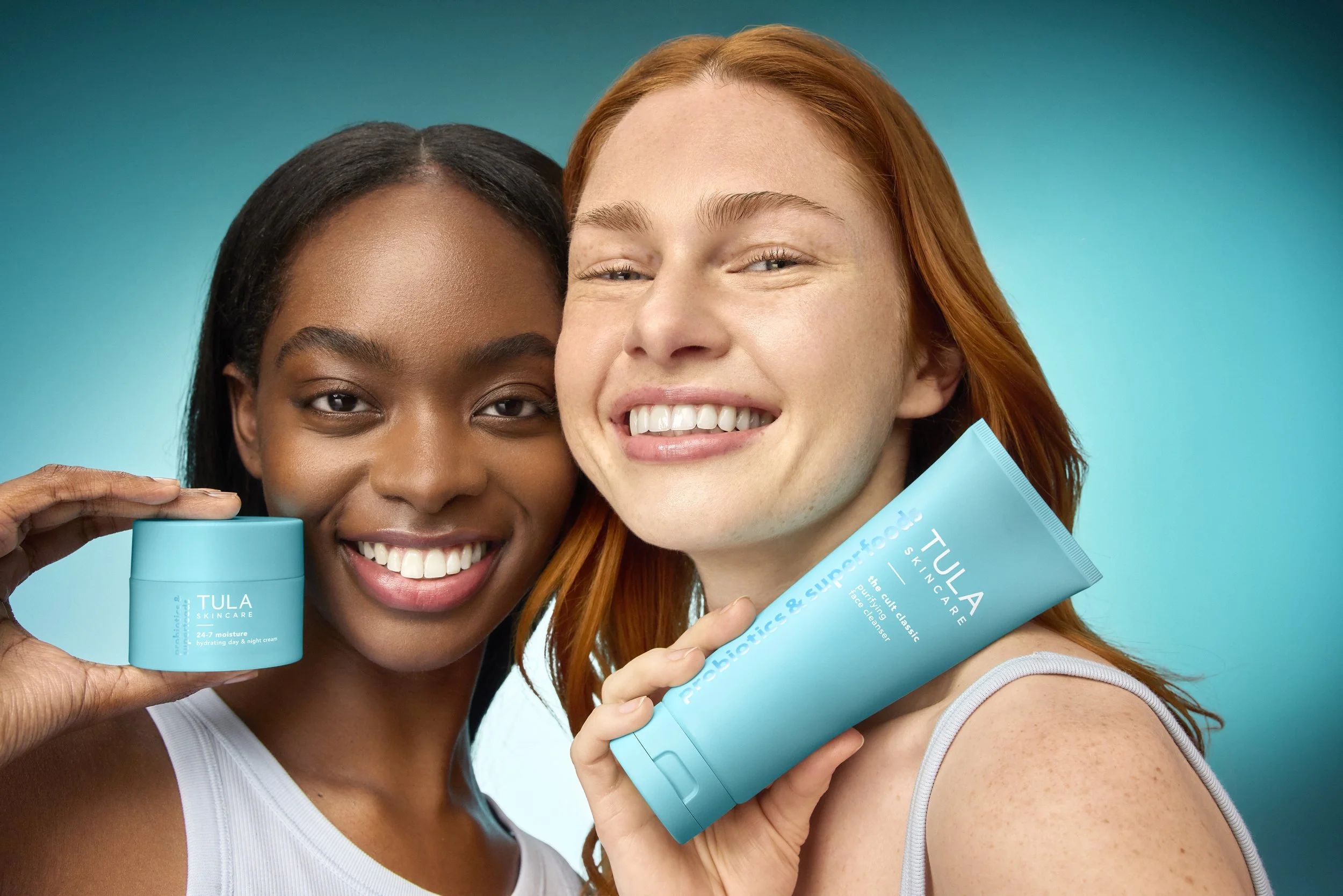 Two women smiling and holding skincare products from TULA Skincare against a bright teal background.