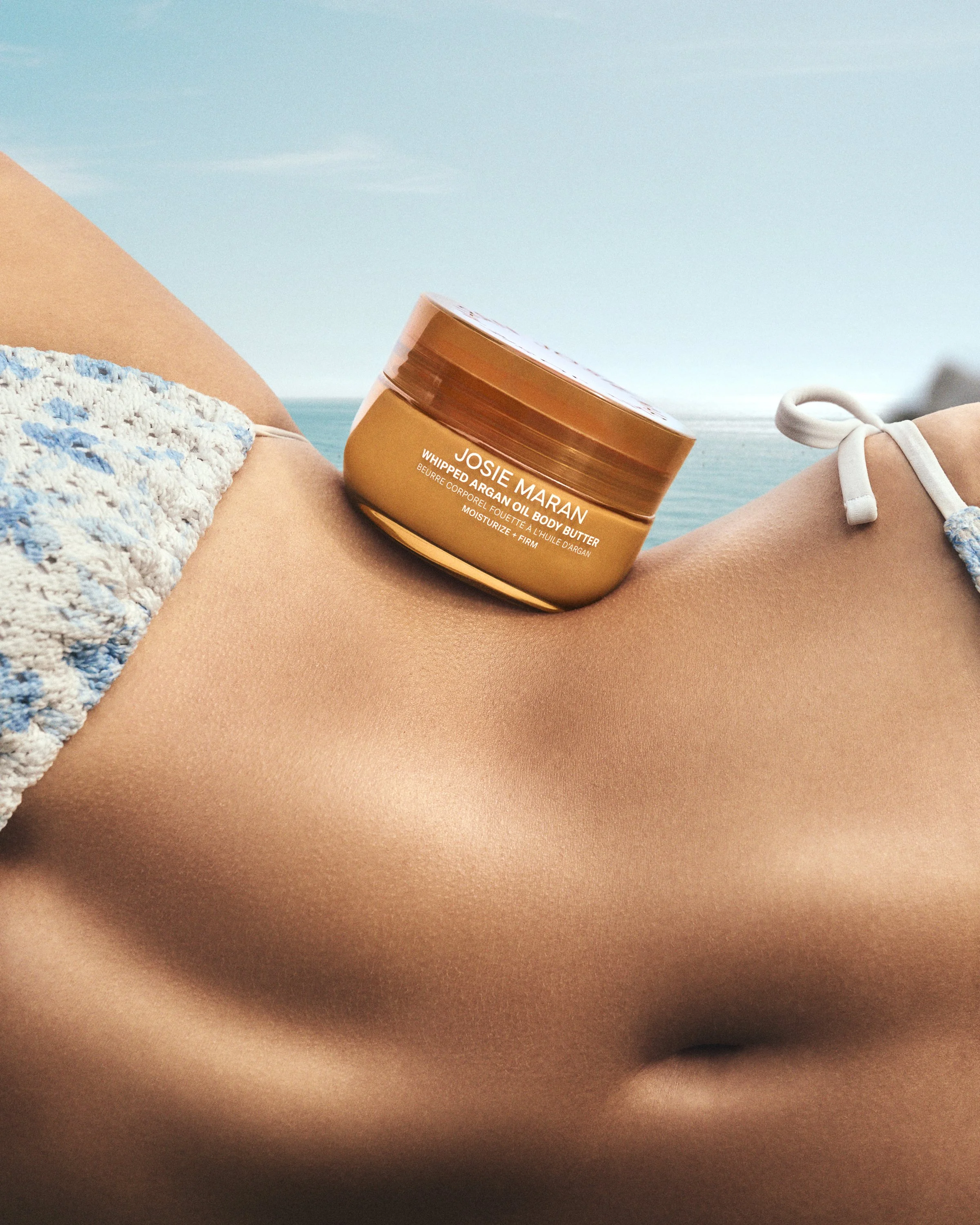 Close-up of a person's body with a jar of Josie Maran whipped argan oil body butter resting on the skin, with ocean and sky in the background.