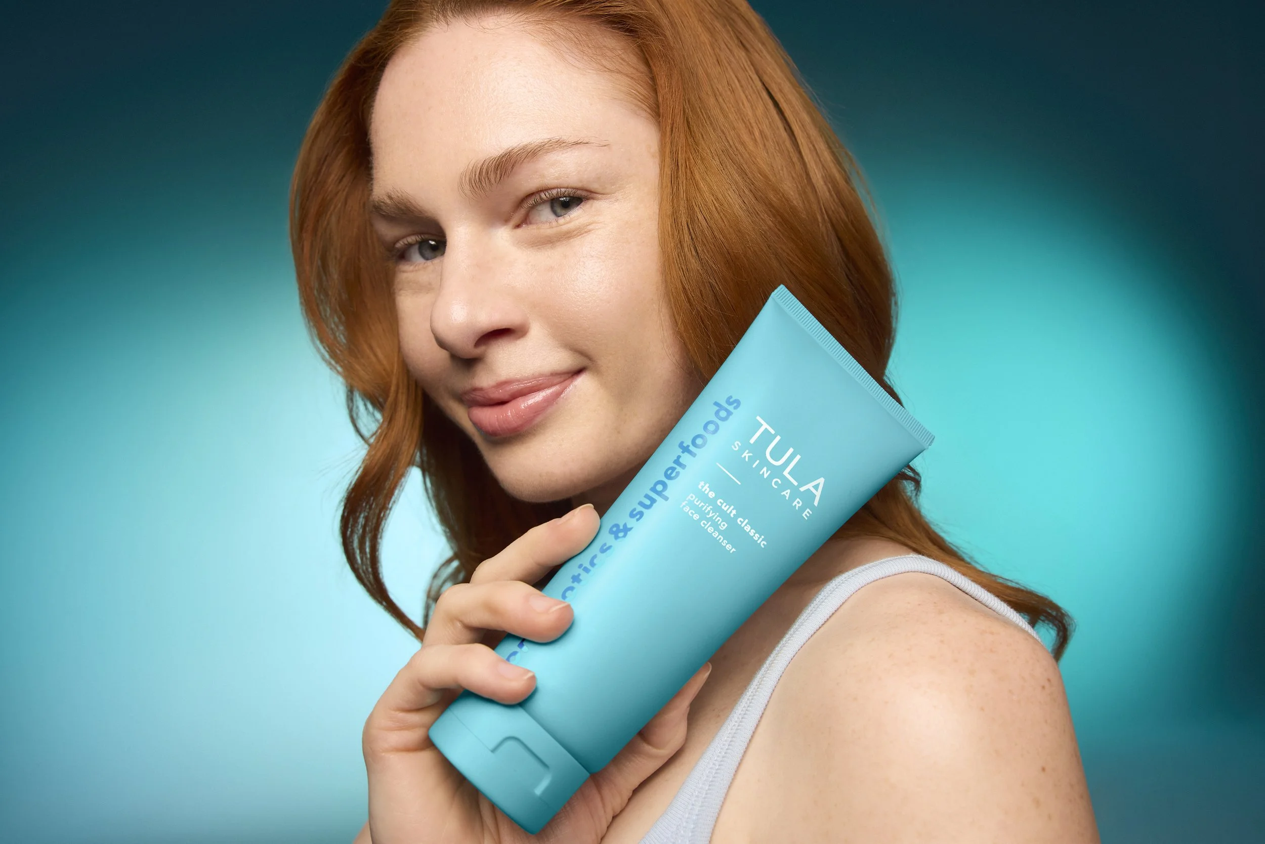 A young woman with red hair holding a turquoise tube of face cleanser labeled 'TULA SKINCARE' against a teal gradient background, smiling slightly.