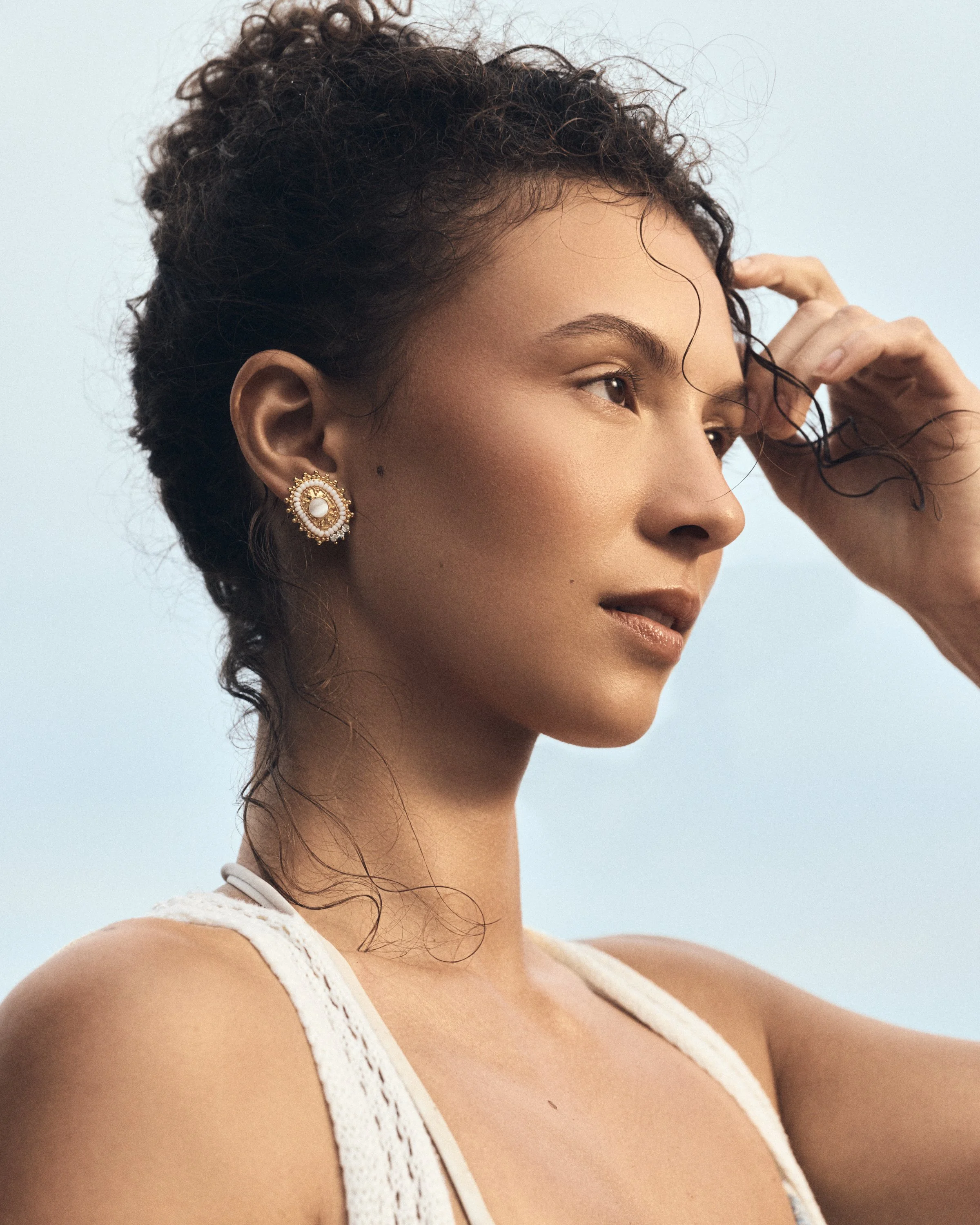 Close-up side profile of a woman with curly hair, gold and pearl earring, wearing a white lace top, against a soft blue background.