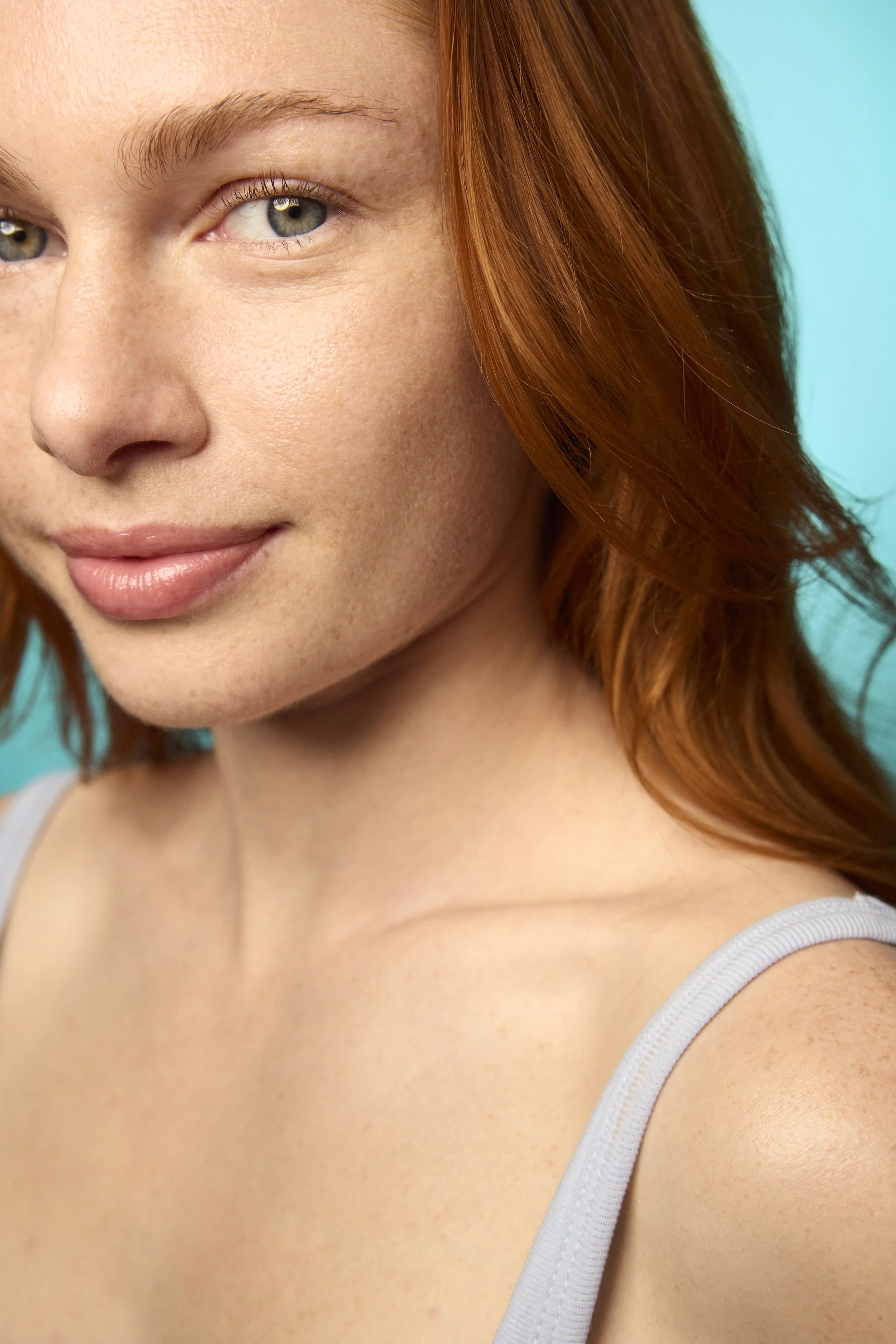 Close-up of a woman's face with red hair, light skin, and wearing a white tank top, set against a teal background.