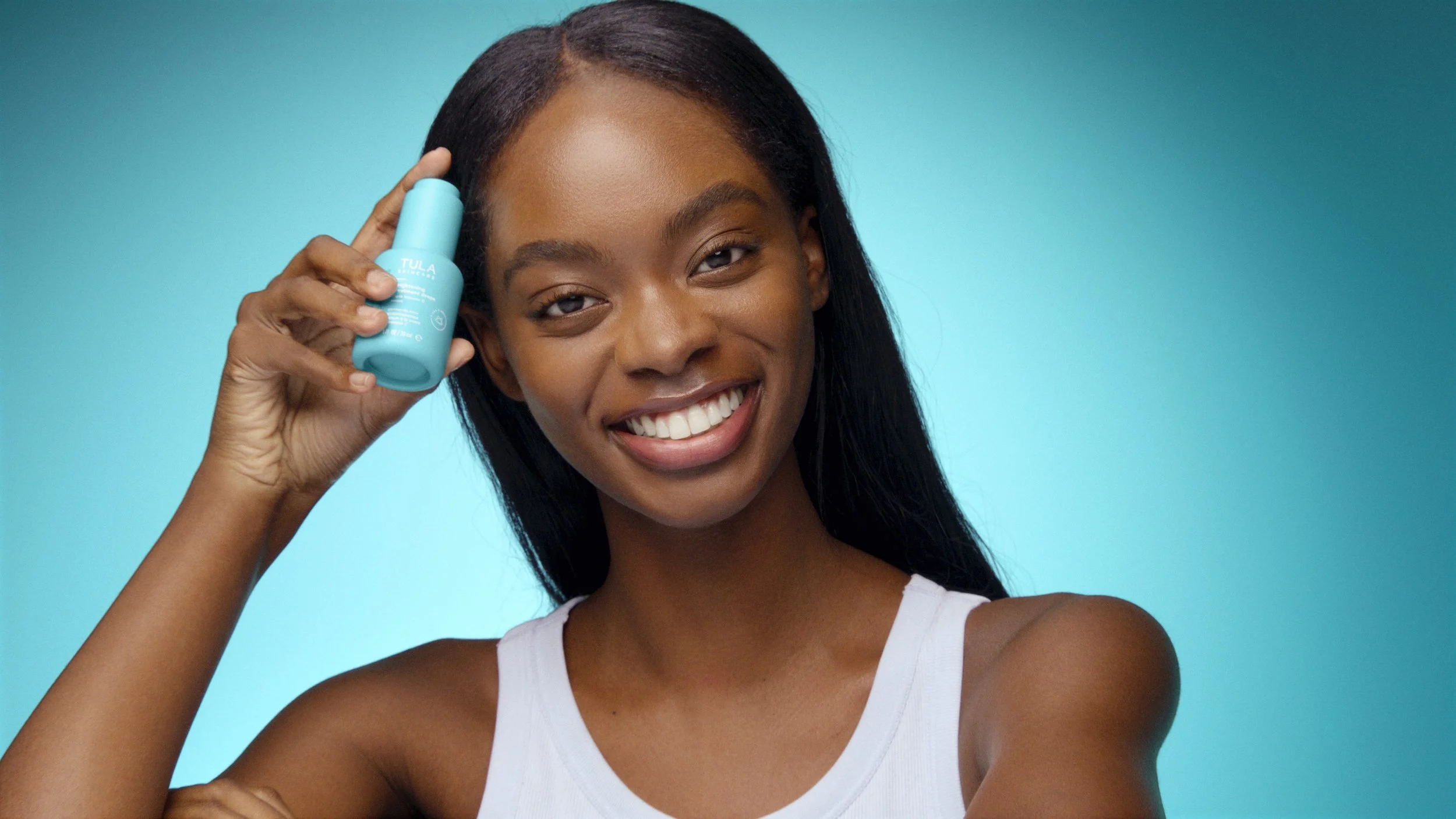 A young woman with long black hair smiling while holding a blue skincare product near her temple against a blue background.