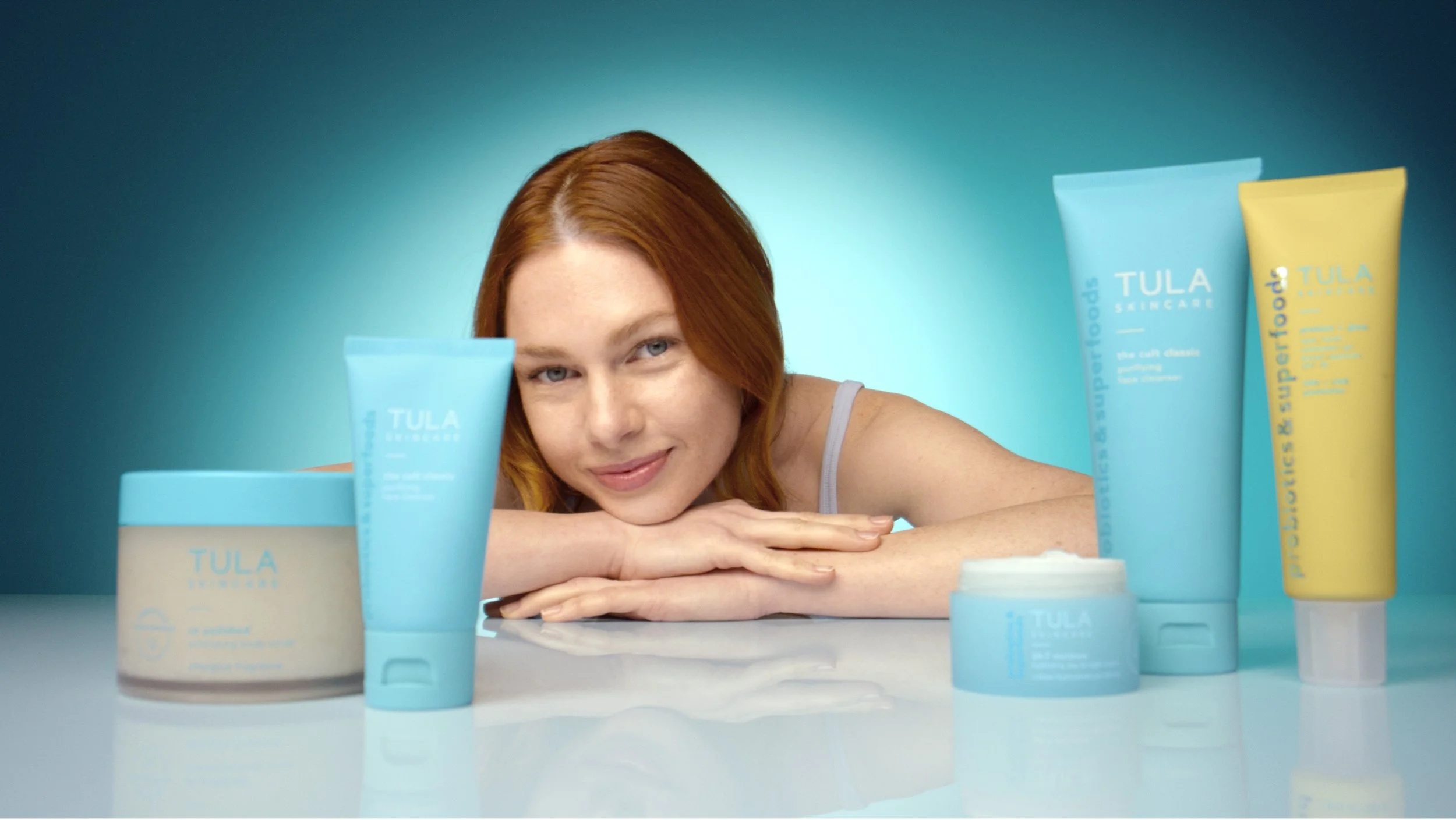 A woman with red hair lies resting her head on her folded arms in front of a display of TULA skincare products, which include tubes and jars arranged on a table against a teal background.