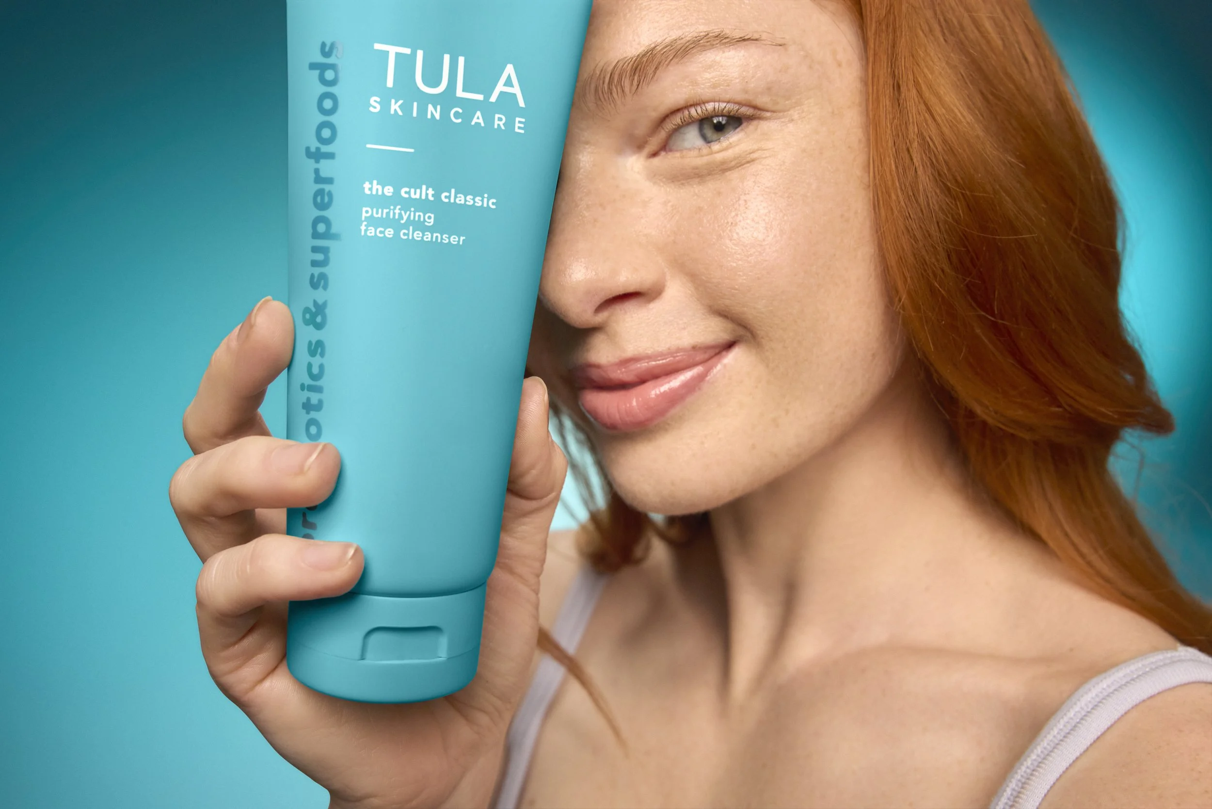 A woman with red hair holding a blue face wash tube with her right hand, partially covering her face, against a blue background.