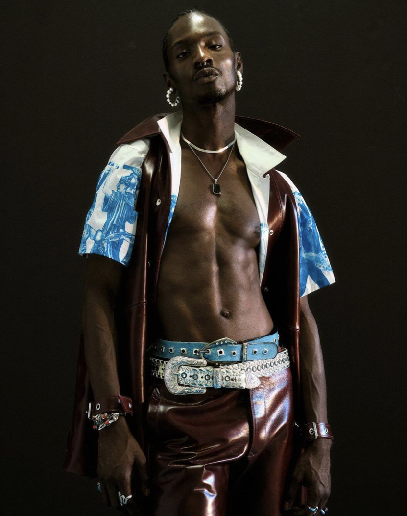 A man with dark skin, styled hair, and piercings, posing against a black background. He is shirtless, wearing a brown leather vest with a blue and white patterned short-sleeve shirt underneath, and matching leather pants. He has jewelry including ear