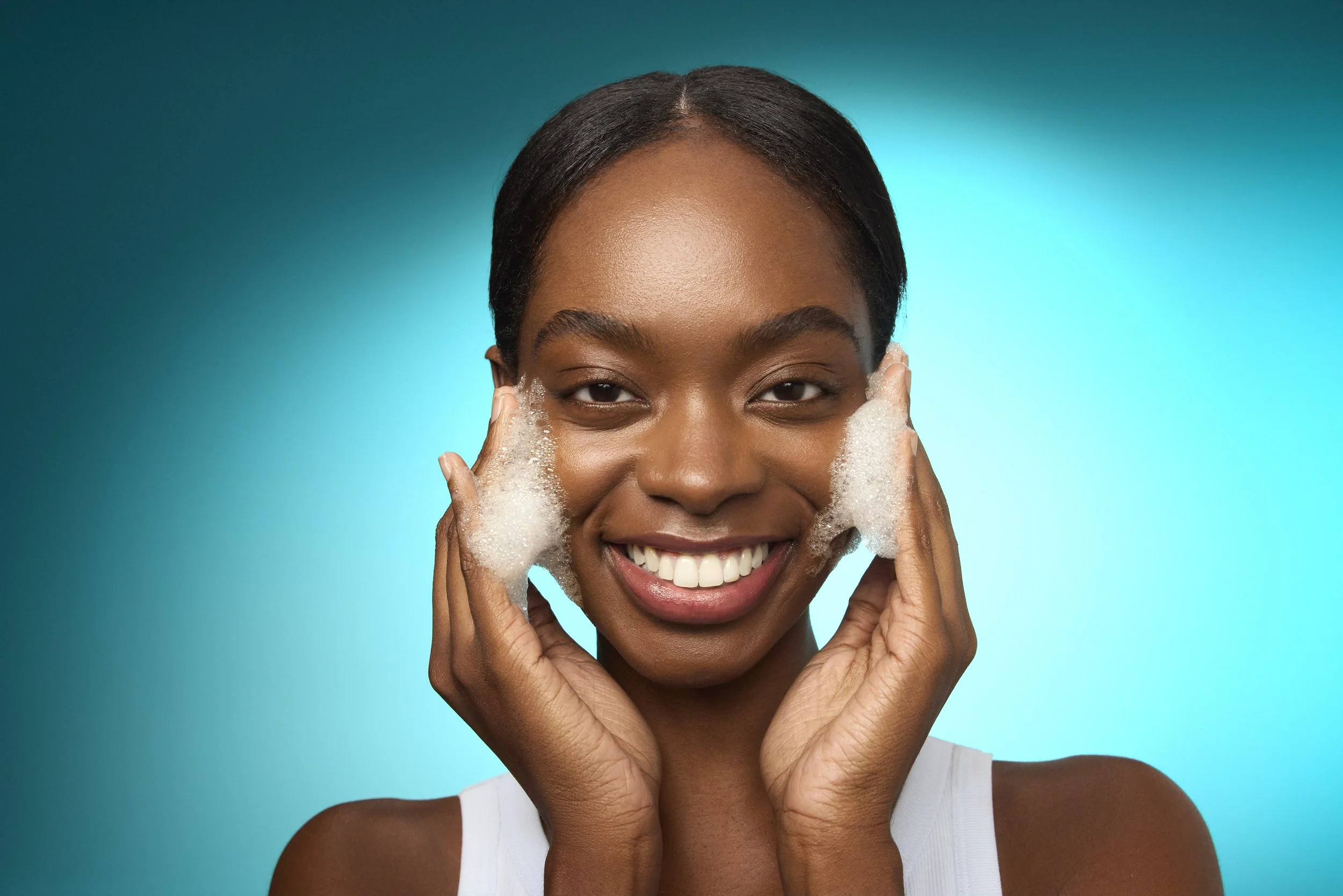 A woman with dark skin smiling while washing her face with soap, surrounded by bubbles, against a bright blue background.