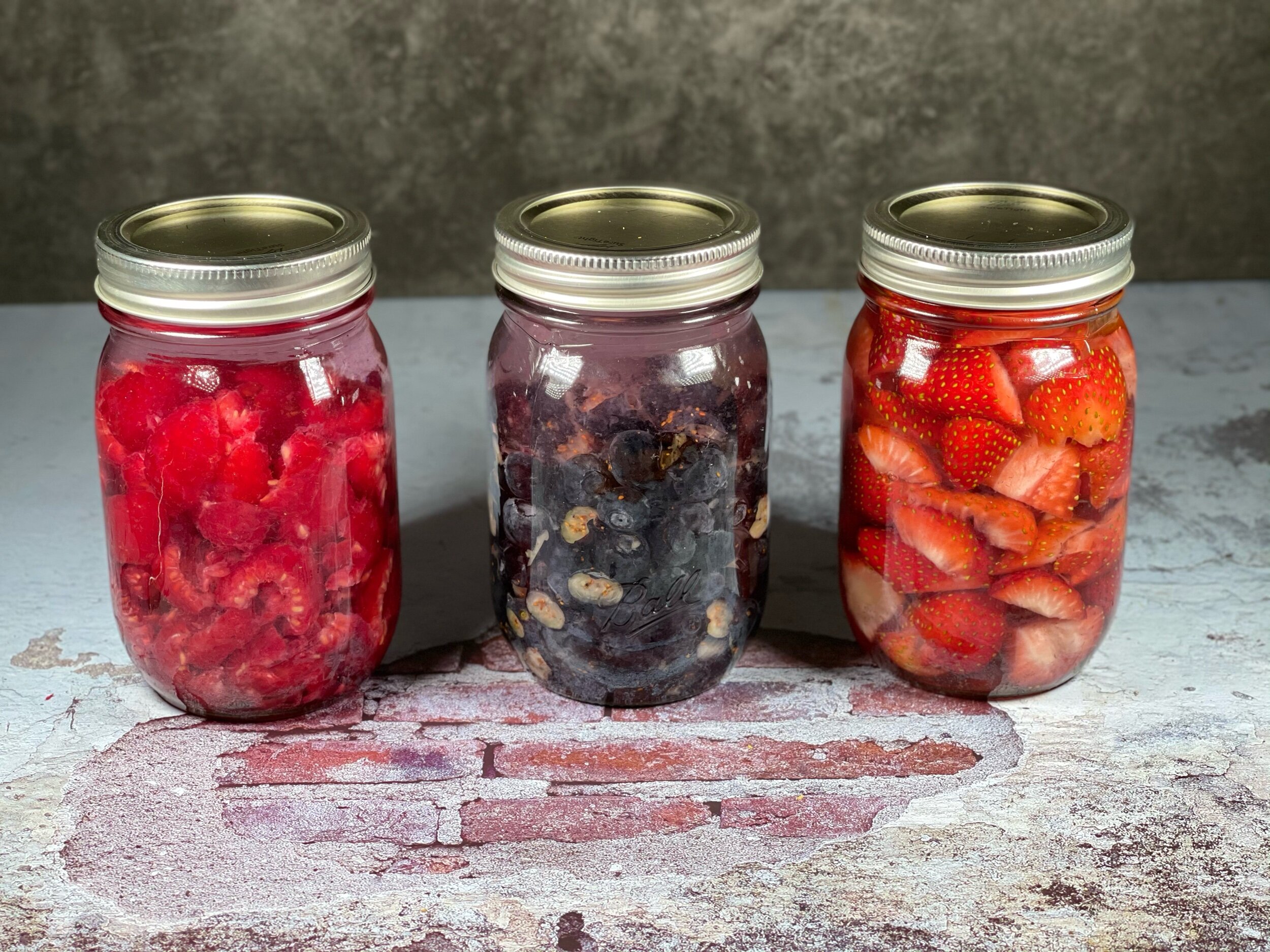  Right to left: mason jars of raspberries, blueberries, and strawberries, all filed with vodka and capped with metal canning lids and rings. 